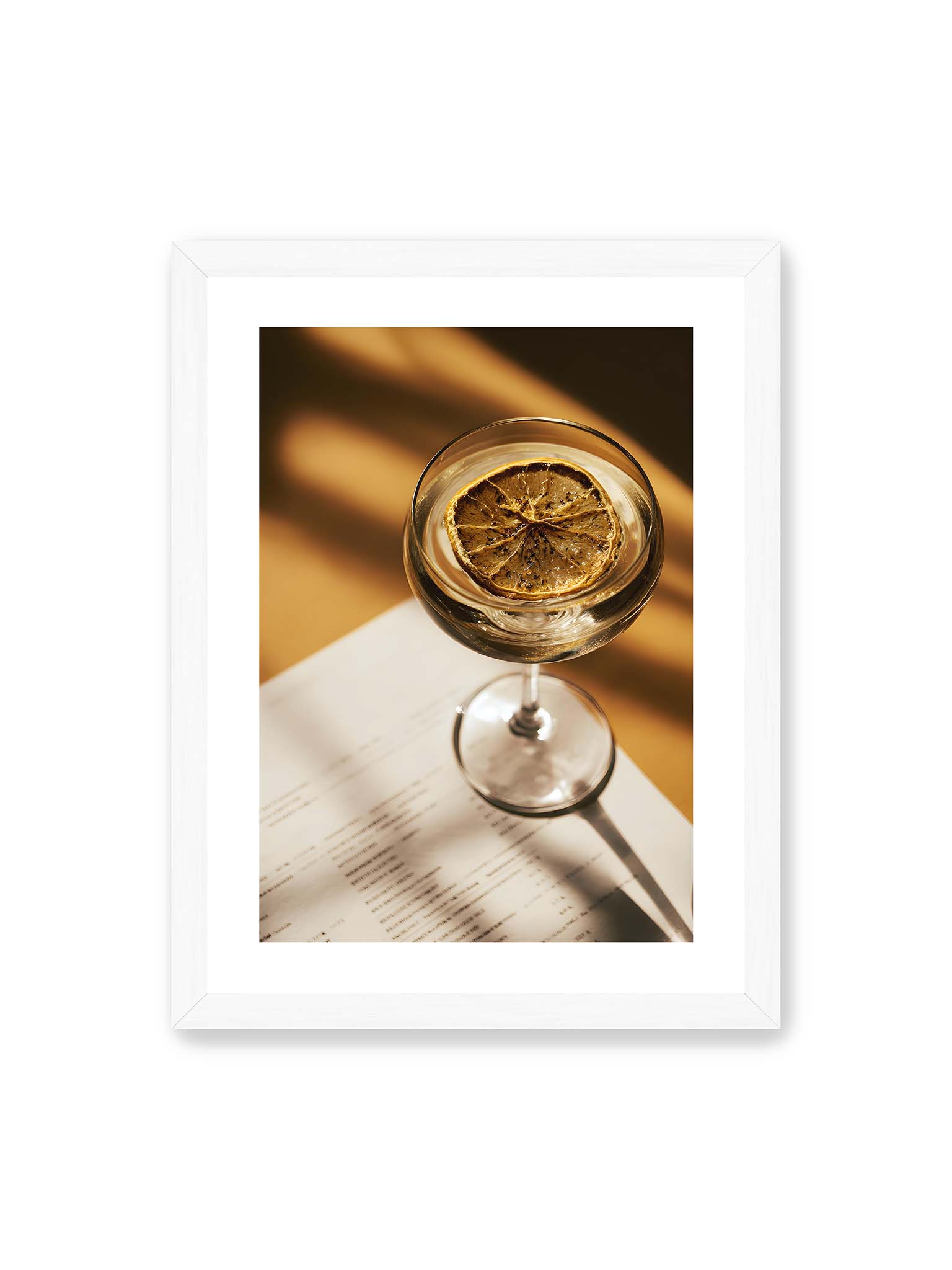 30x40 cm, weißer Galerie-Rand, weißer Rahmen – Cocktailglas mit klarem Getränk und getrockneter Zitrusscheibe von oben fotografiert, warmes Licht und Schatten auf Tisch und Papier.