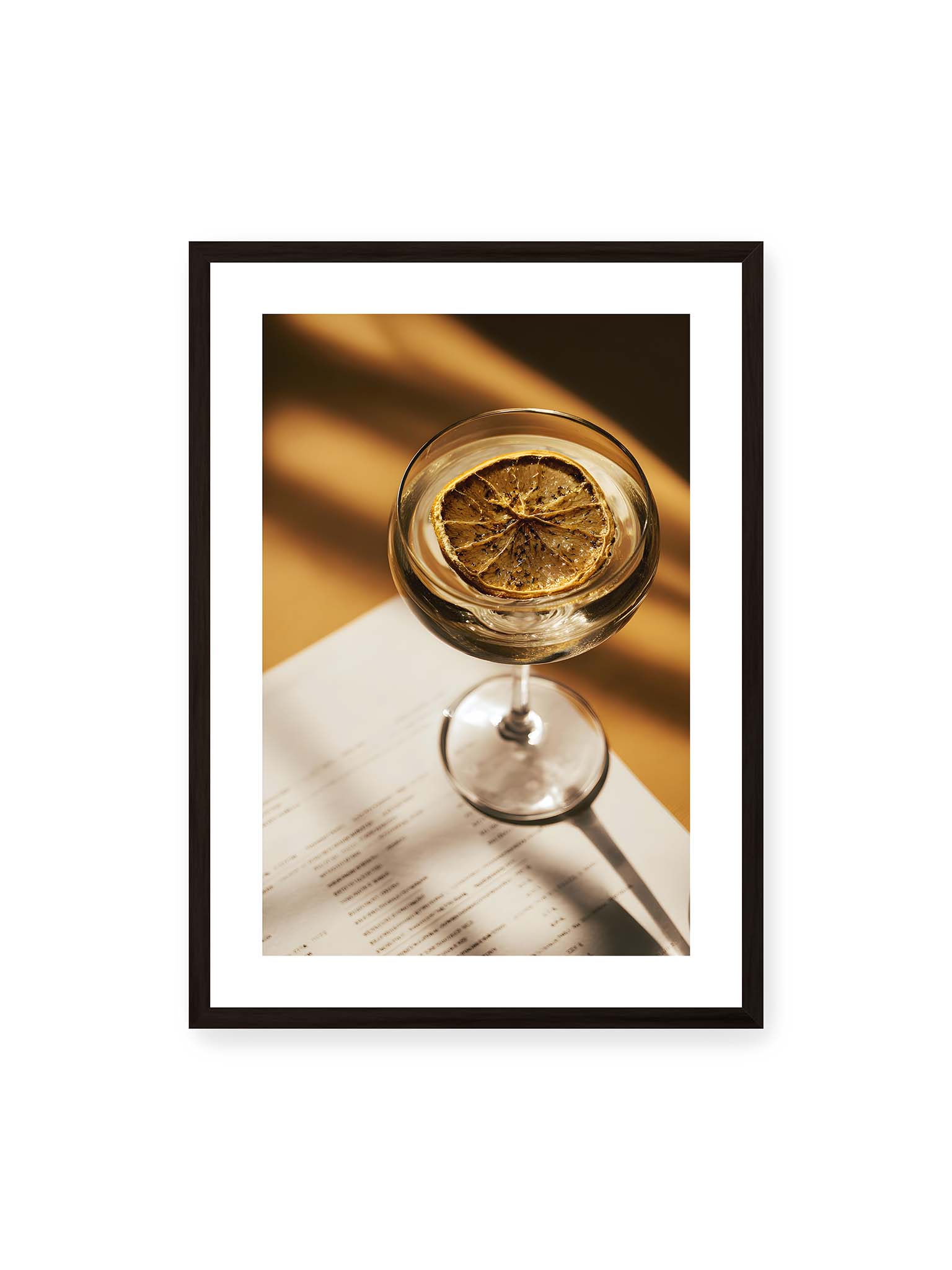 50x70 cm, weißer Galerie-Rand, schwarzer Rahmen – Cocktailglas mit klarem Getränk und getrockneter Zitrusscheibe von oben fotografiert, warmes Licht und Schatten auf Tisch und Papier.