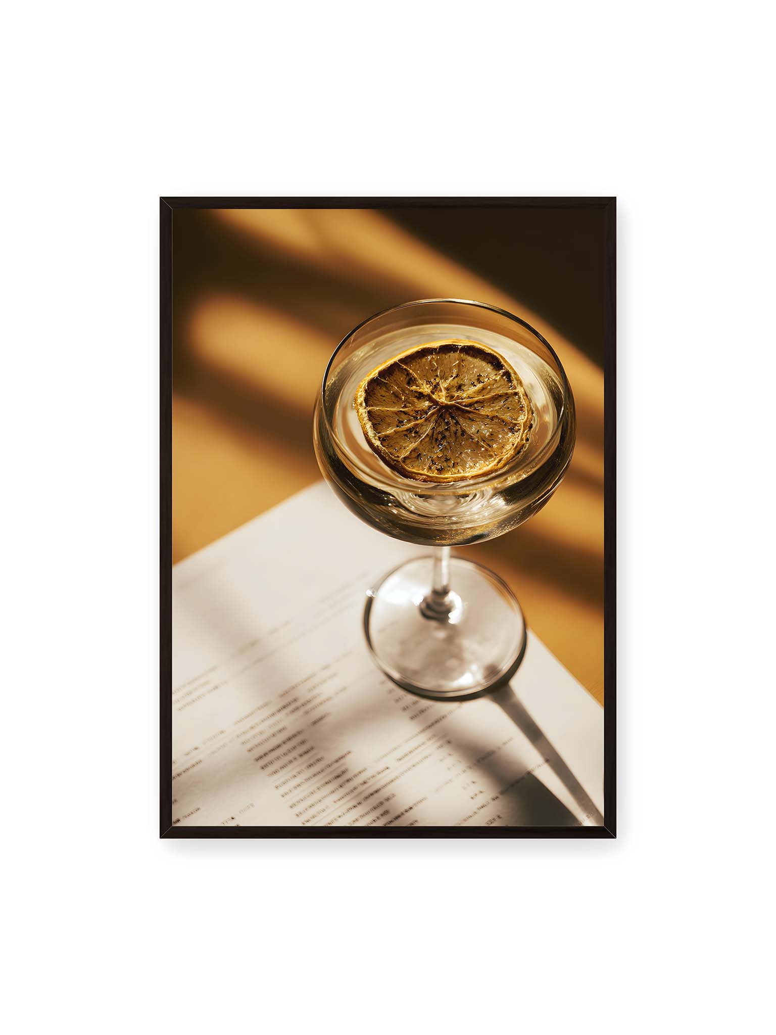70x100 cm, schwarzer Rahmen – Cocktailglas mit klarem Getränk und getrockneter Zitrusscheibe von oben fotografiert, warmes Licht und Schatten auf Tisch und Papier.