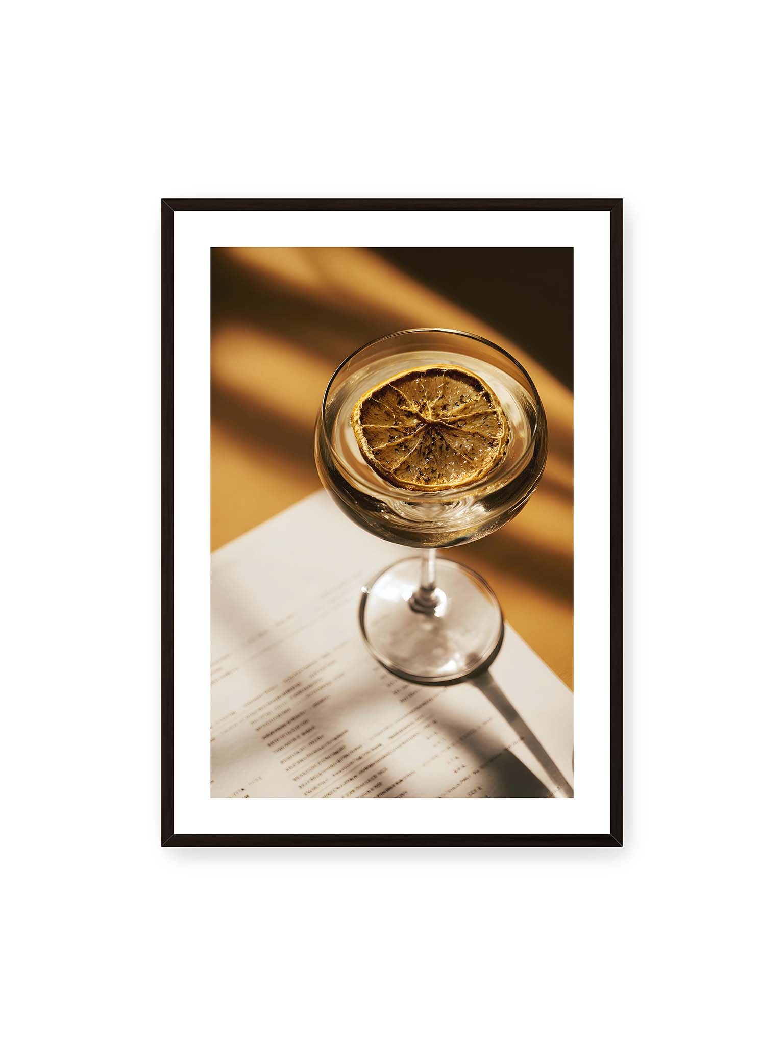 70x100 cm, weißer Galerie-Rand, schwarzer Rahmen – Cocktailglas mit klarem Getränk und getrockneter Zitrusscheibe von oben fotografiert, warmes Licht und Schatten auf Tisch und Papier.