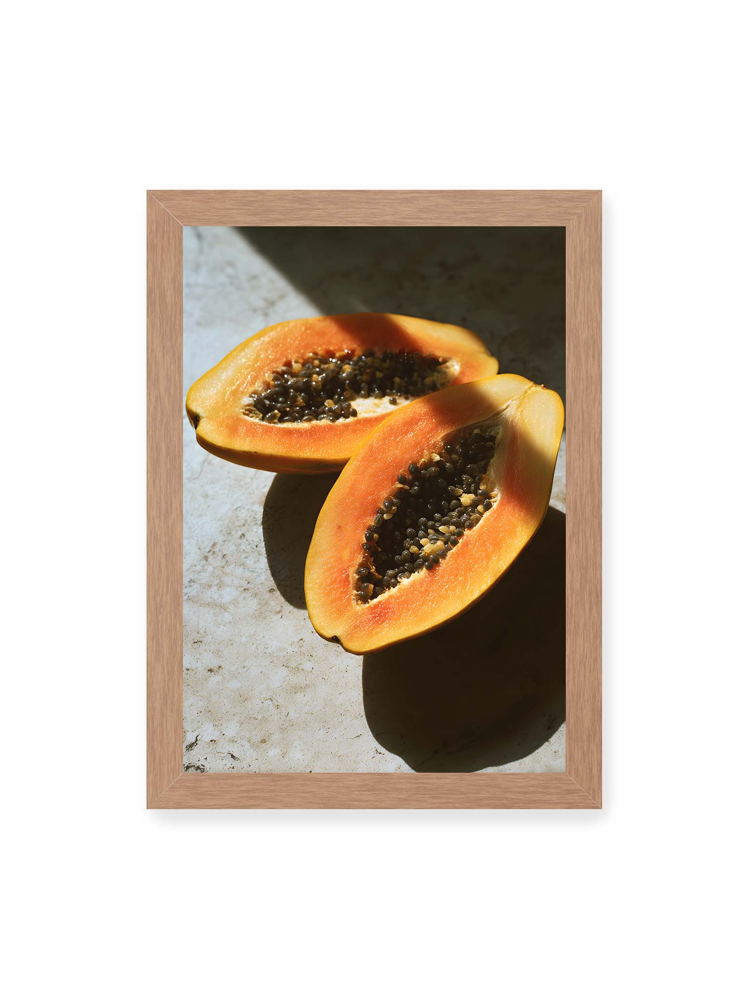 21x30 cm, Eichenrahmen – Zwei halbierte Papayas mit dunklen Samen auf heller Oberfläche, beleuchtet von warmem Licht mit starken Schatten.