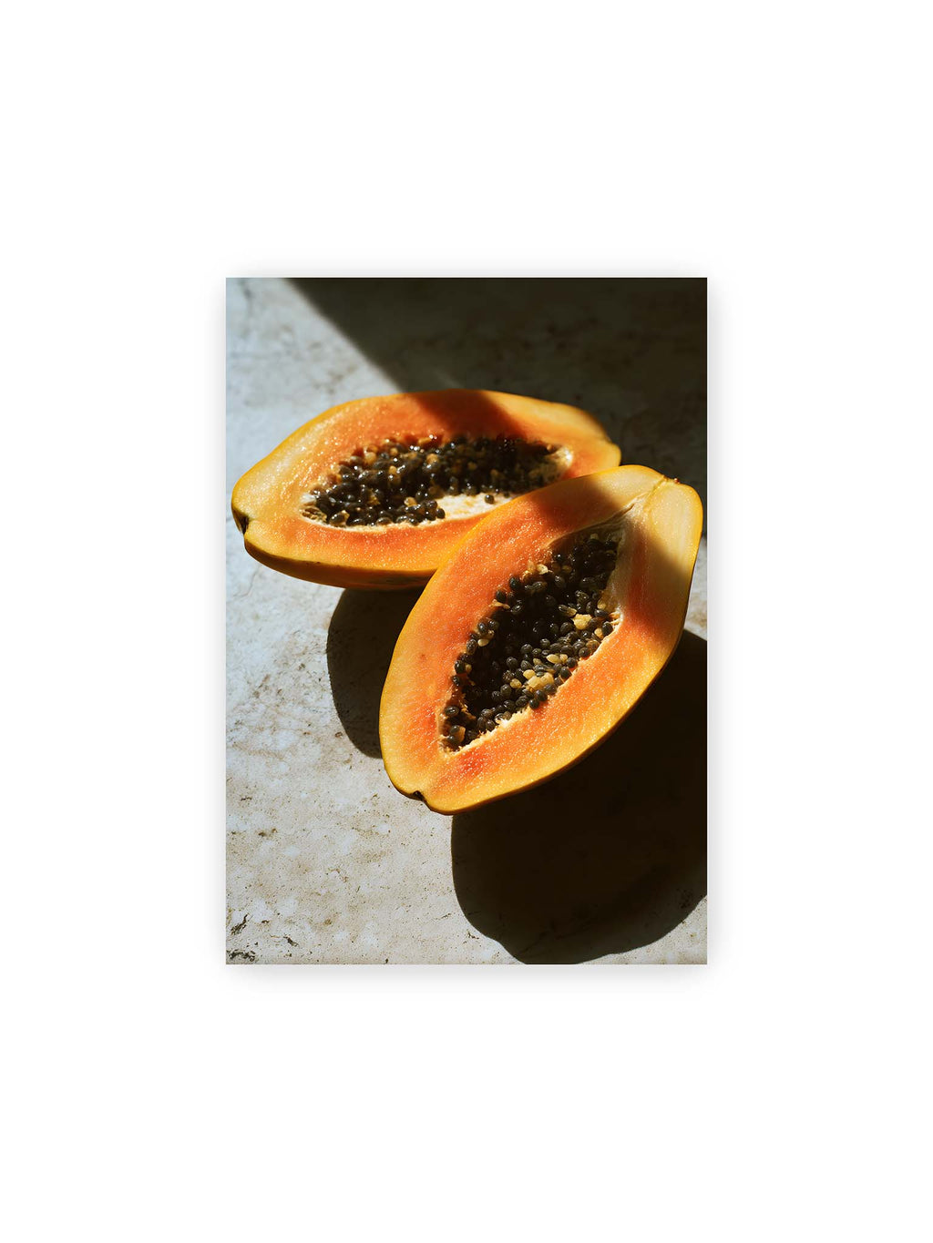 21x30 cm – Zwei halbierte Papayas mit dunklen Samen auf heller Oberfläche, beleuchtet von warmem Licht mit starken Schatten.