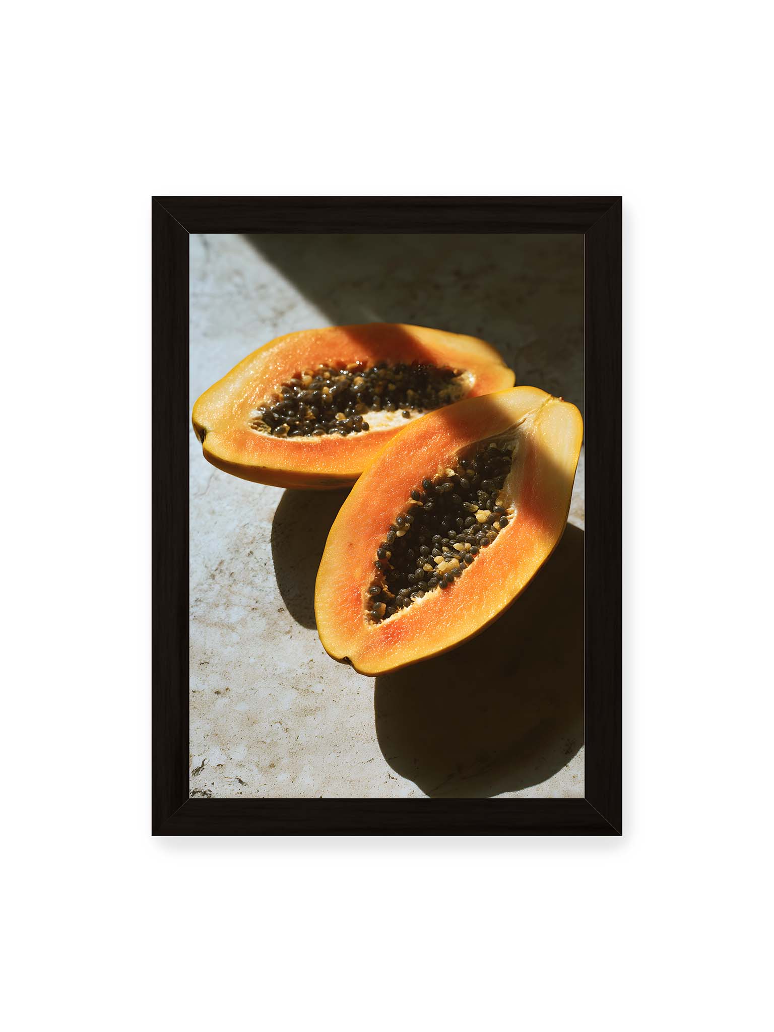 21x30 cm, schwarzer Rahmen – Zwei halbierte Papayas mit dunklen Samen auf heller Oberfläche, beleuchtet von warmem Licht mit starken Schatten.