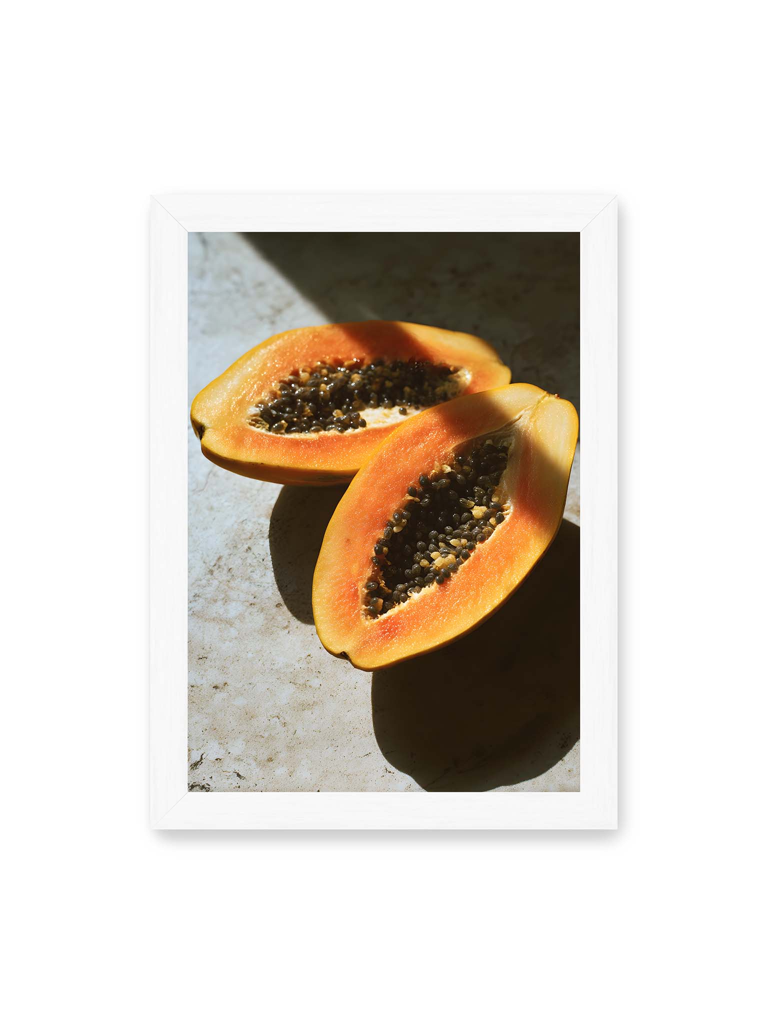 21x30 cm, weißer Rahmen – Zwei halbierte Papayas mit dunklen Samen auf heller Oberfläche, beleuchtet von warmem Licht mit starken Schatten.