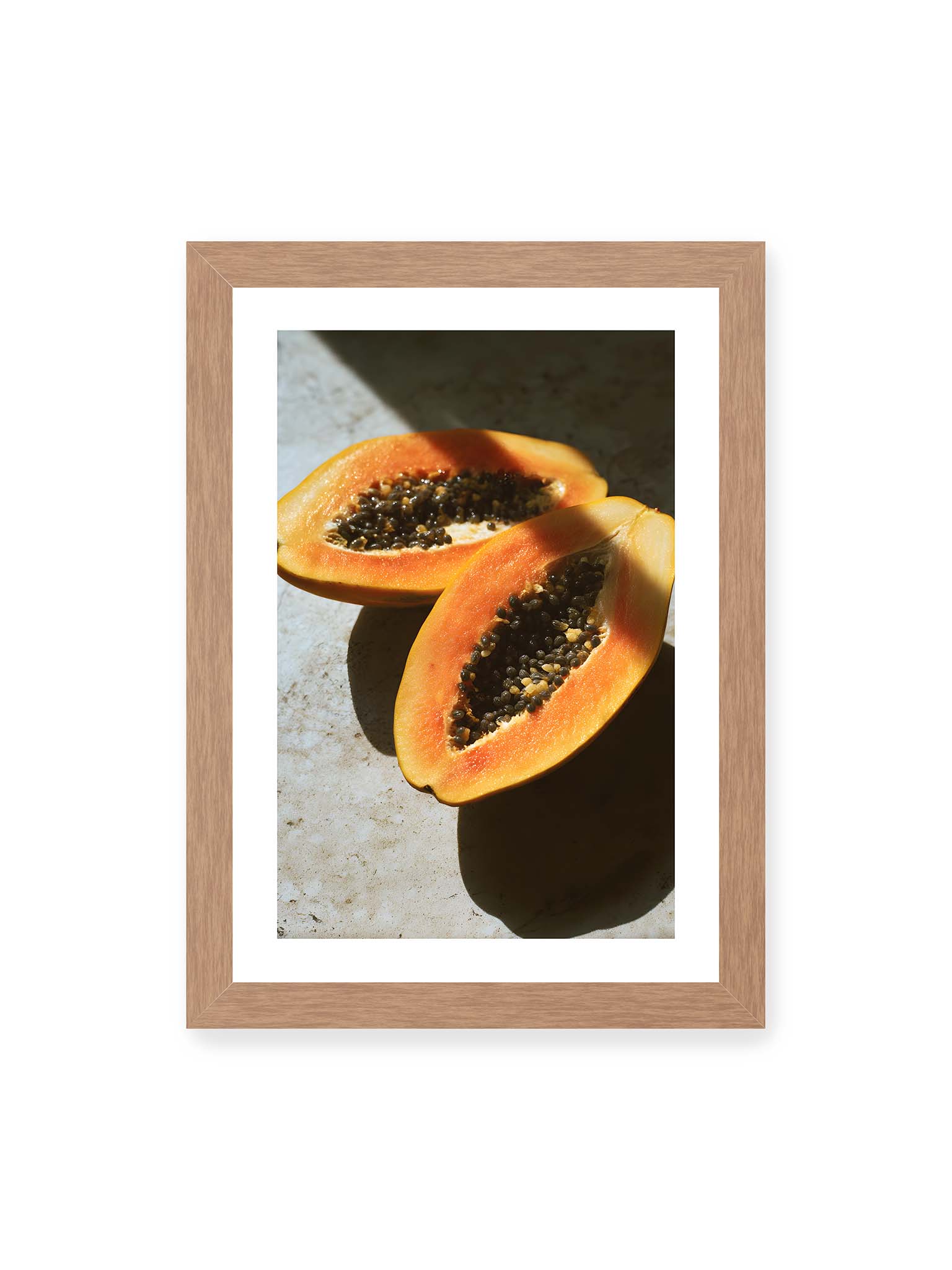 21x30 cm, weißer Galerie-Rand, Eichenrahmen – Zwei halbierte Papayas mit dunklen Samen auf heller Oberfläche, beleuchtet von warmem Licht mit starken Schatten.