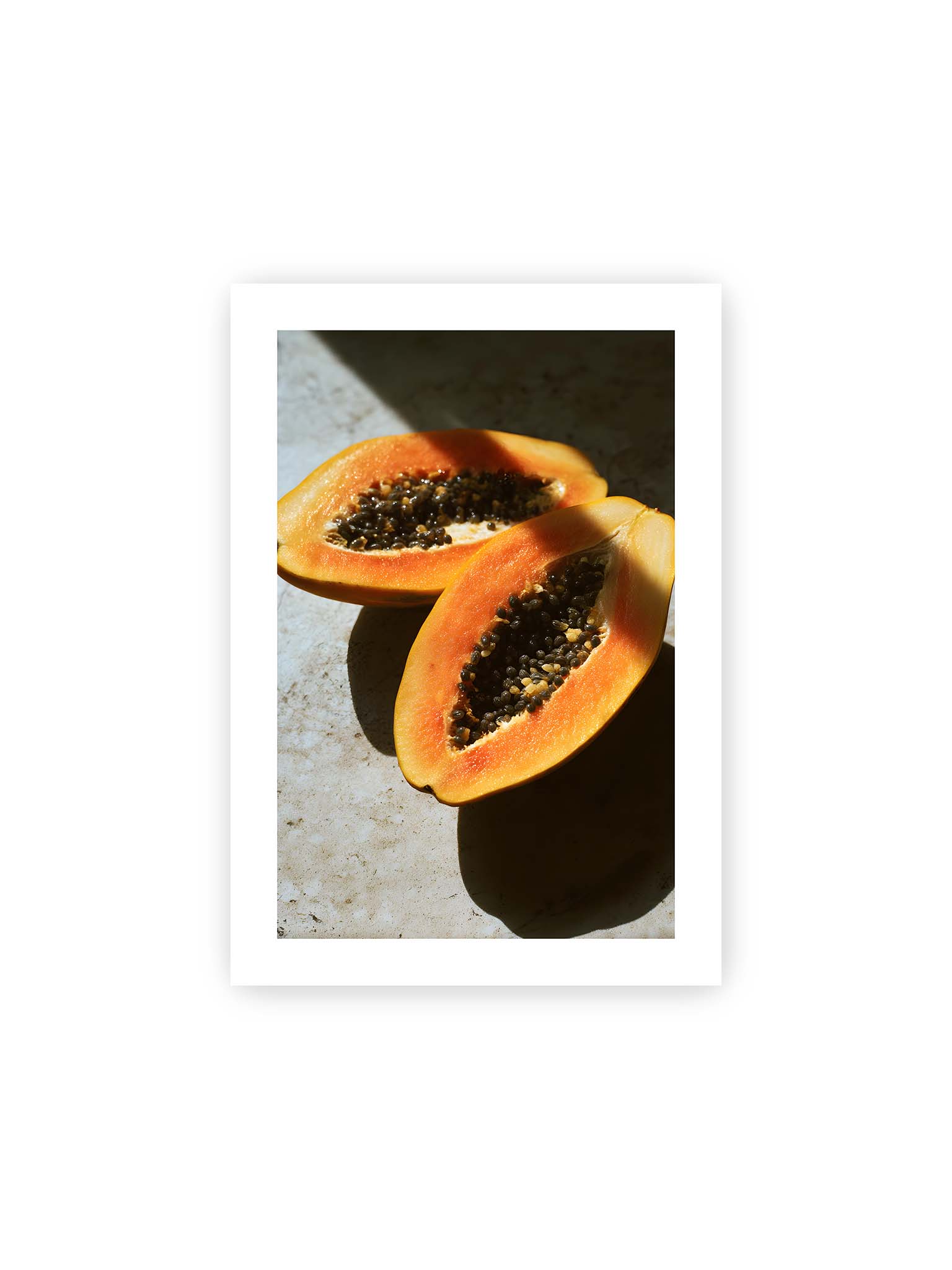 21x30 cm, weißer Galerie-Rand – Zwei halbierte Papayas mit dunklen Samen auf heller Oberfläche, beleuchtet von warmem Licht mit starken Schatten.