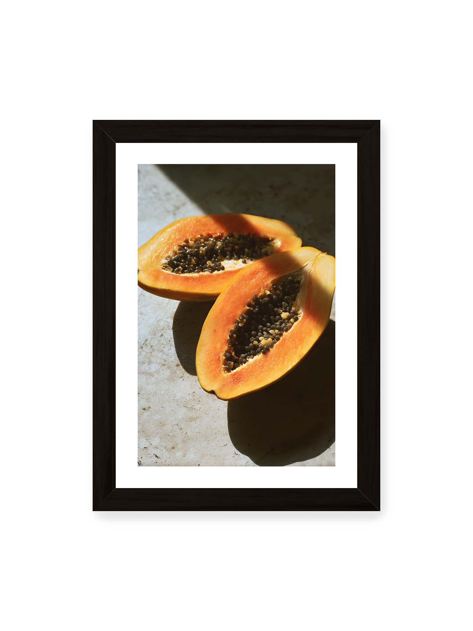 21x30 cm, weißer Galerie-Rand, schwarzer Rahmen – Zwei halbierte Papayas mit dunklen Samen auf heller Oberfläche, beleuchtet von warmem Licht mit starken Schatten.