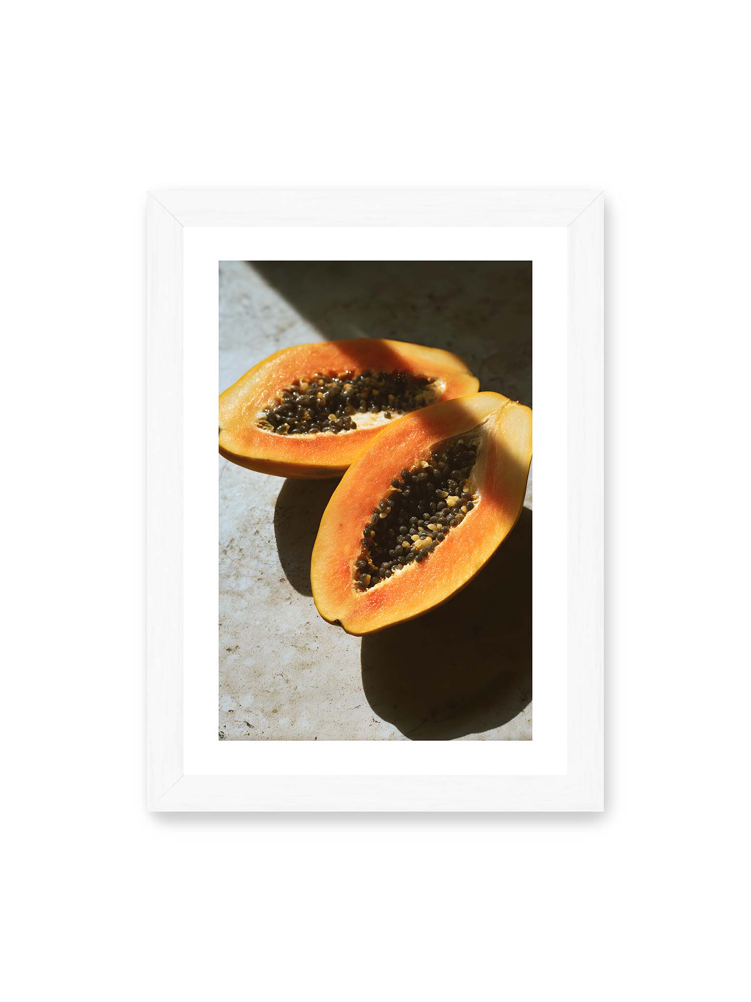21x30 cm, weißer Galerie-Rand, weißer Rahmen – Zwei halbierte Papayas mit dunklen Samen auf heller Oberfläche, beleuchtet von warmem Licht mit starken Schatten.