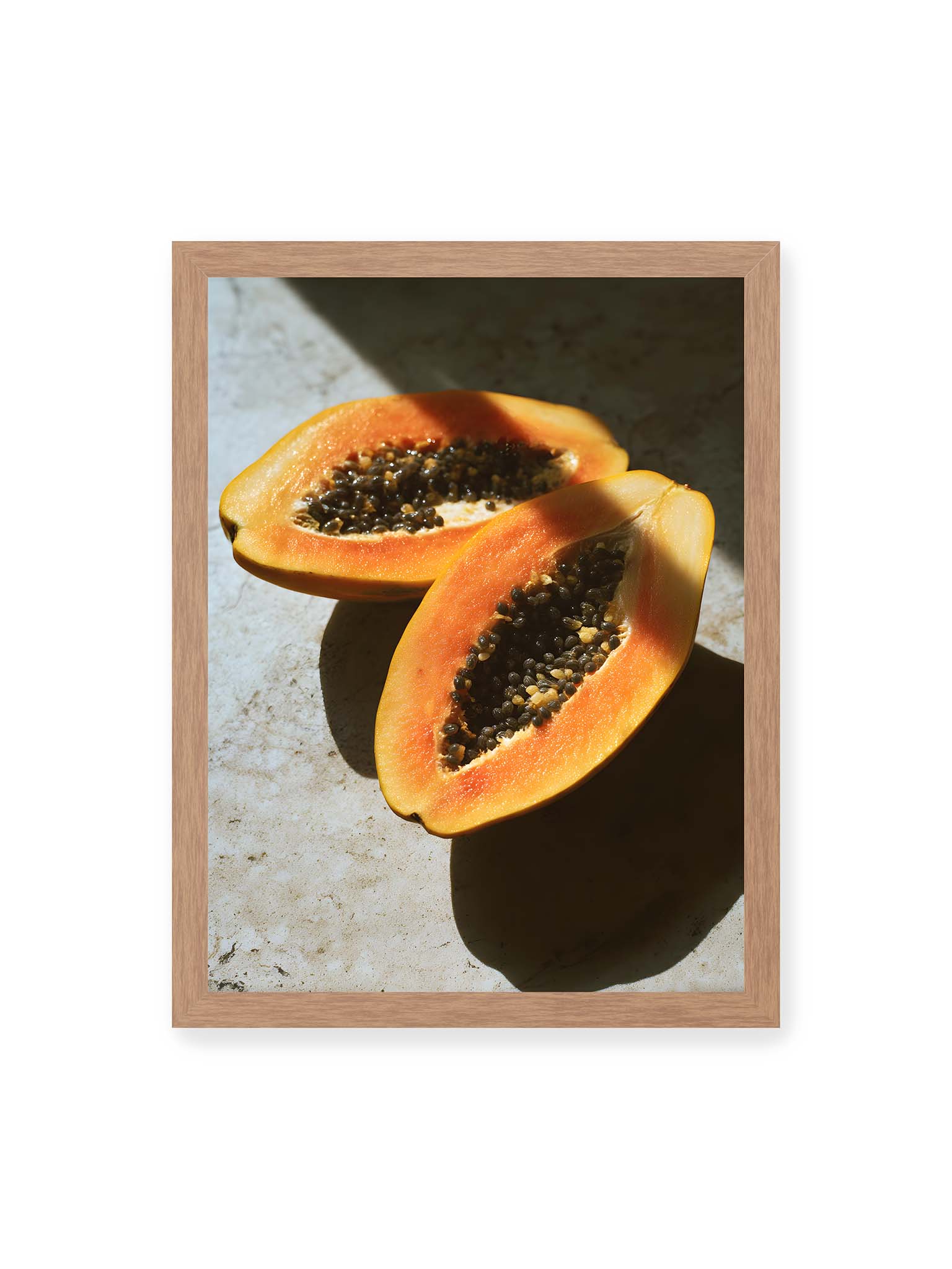 30x40 cm, Eichenrahmen – Zwei halbierte Papayas mit dunklen Samen auf heller Oberfläche, beleuchtet von warmem Licht mit starken Schatten.