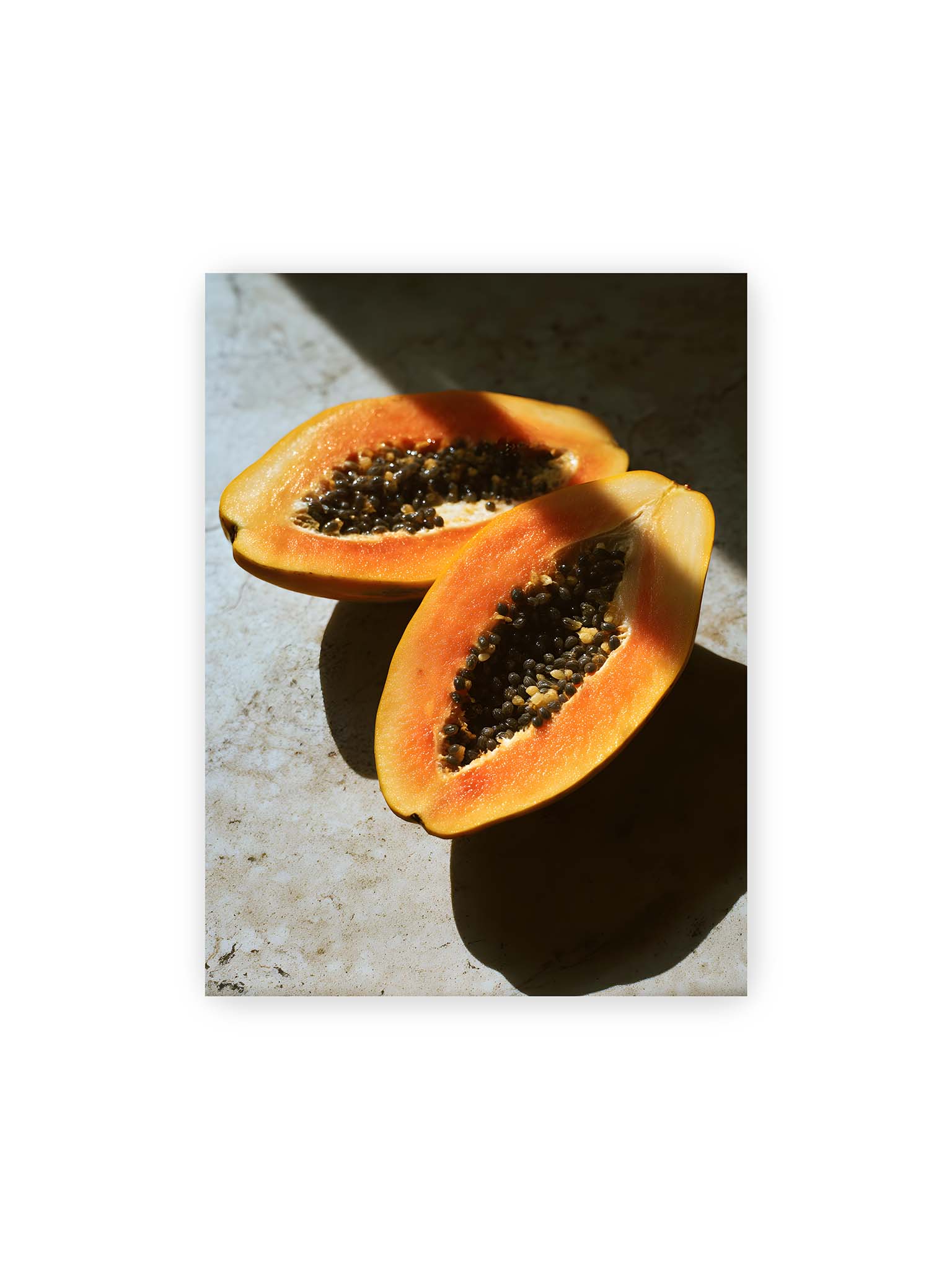 30x40 cm – Zwei halbierte Papayas mit dunklen Samen auf heller Oberfläche, beleuchtet von warmem Licht mit starken Schatten.