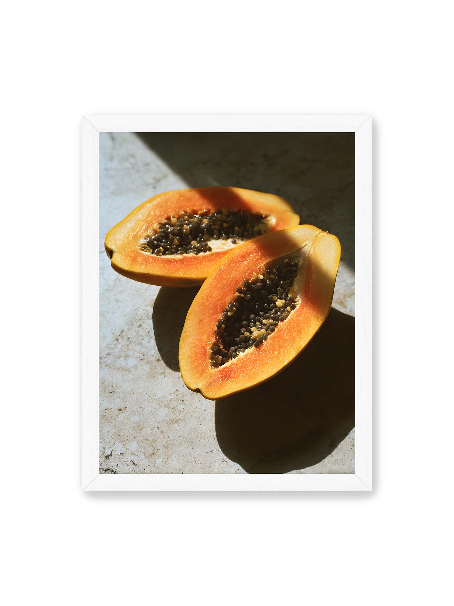 30x40 cm, weißer Rahmen – Zwei halbierte Papayas mit dunklen Samen auf heller Oberfläche, beleuchtet von warmem Licht mit starken Schatten.