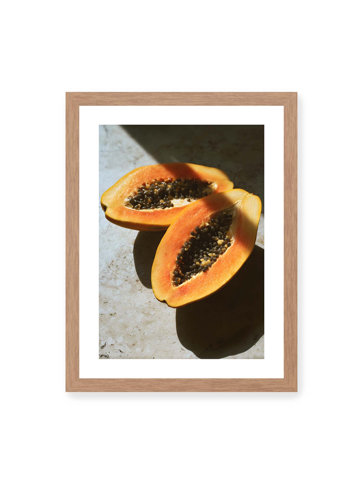 30x40 cm, weißer Galerie-Rand, Eichenrahmen – Zwei halbierte Papayas mit dunklen Samen auf heller Oberfläche, beleuchtet von warmem Licht mit starken Schatten.