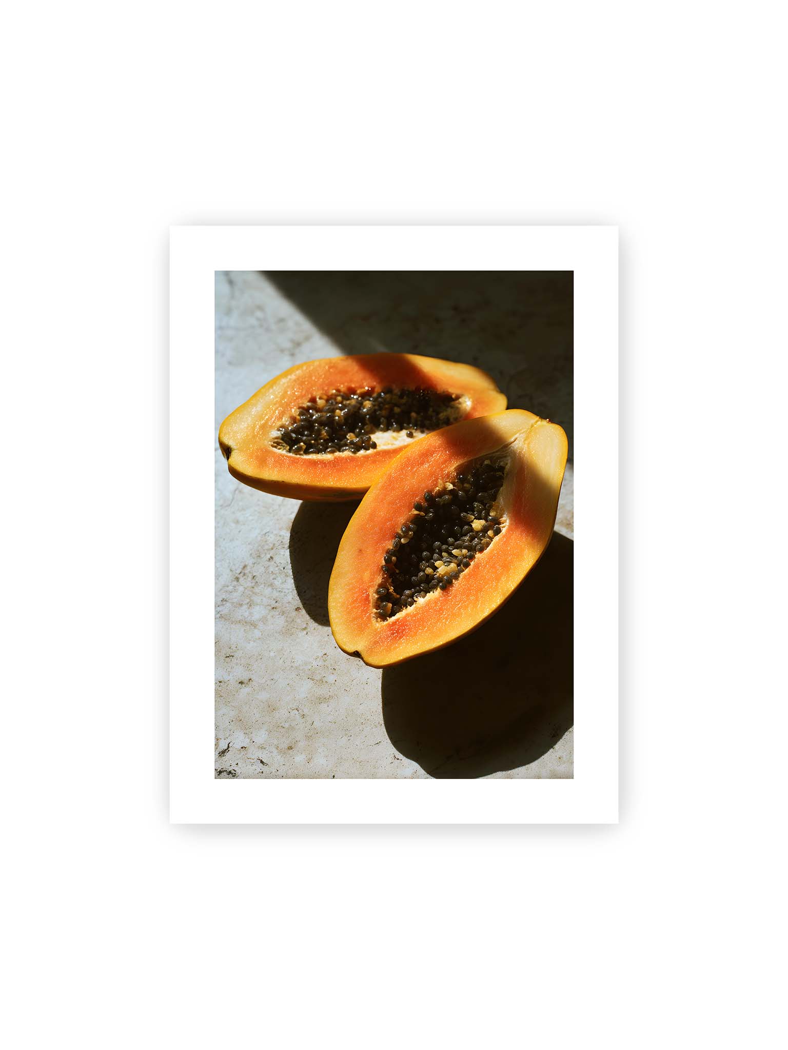30x40 cm, weißer Galerie-Rand – Zwei halbierte Papayas mit dunklen Samen auf heller Oberfläche, beleuchtet von warmem Licht mit starken Schatten.