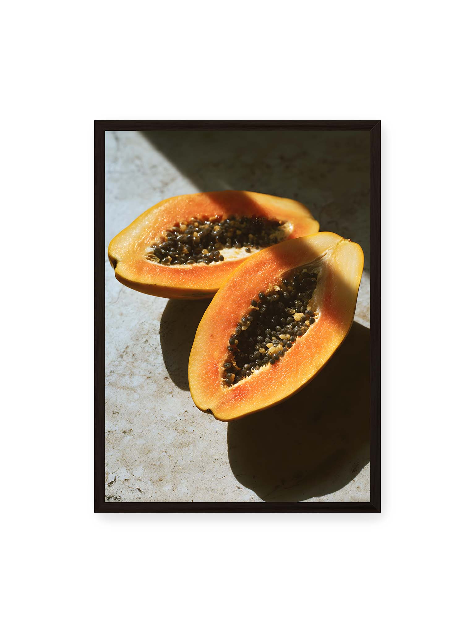 50x70 cm, schwarzer Rahmen – Zwei halbierte Papayas mit dunklen Samen auf heller Oberfläche, beleuchtet von warmem Licht mit starken Schatten.