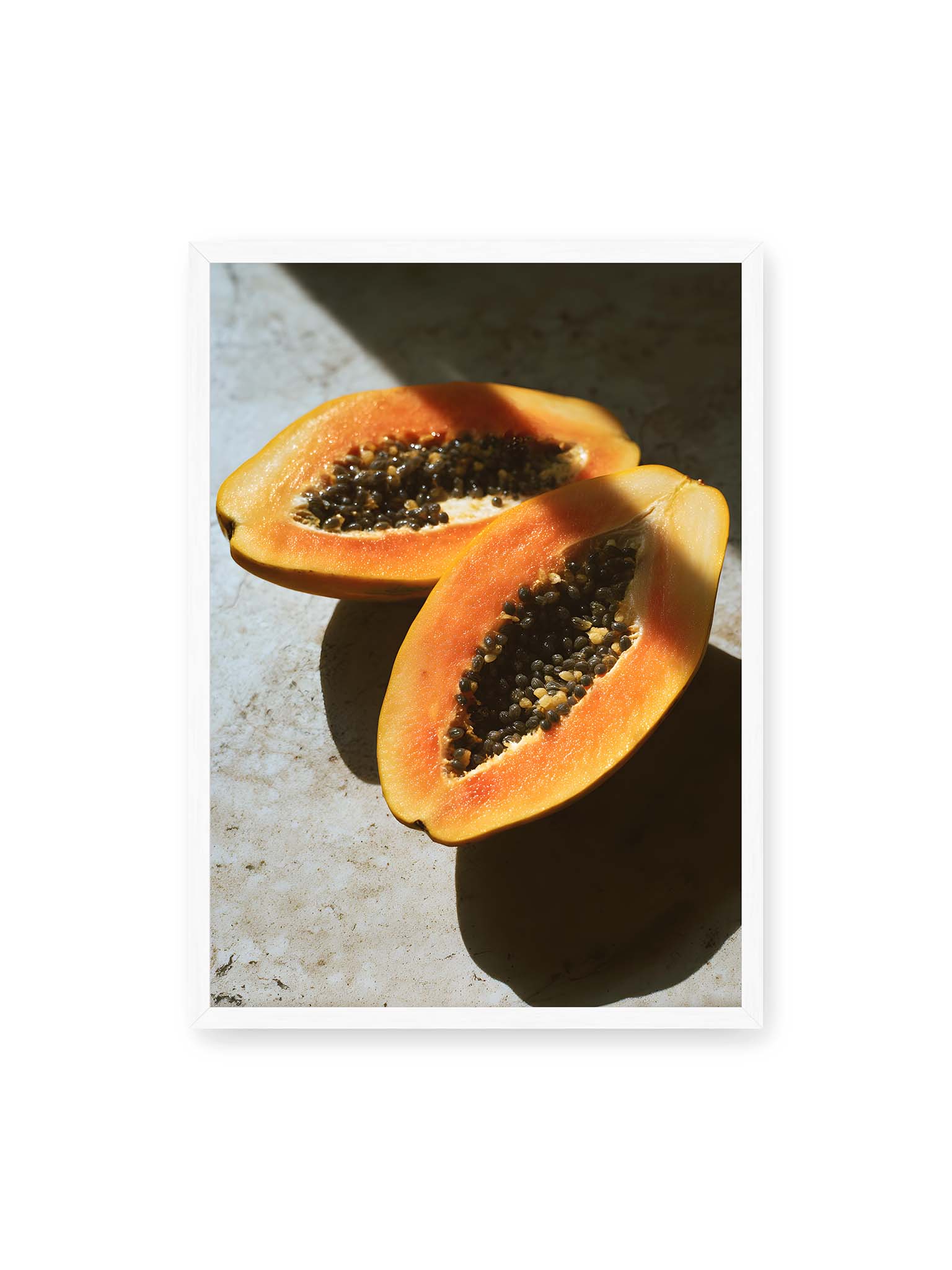 50x70 cm, weißer Rahmen – Zwei halbierte Papayas mit dunklen Samen auf heller Oberfläche, beleuchtet von warmem Licht mit starken Schatten.