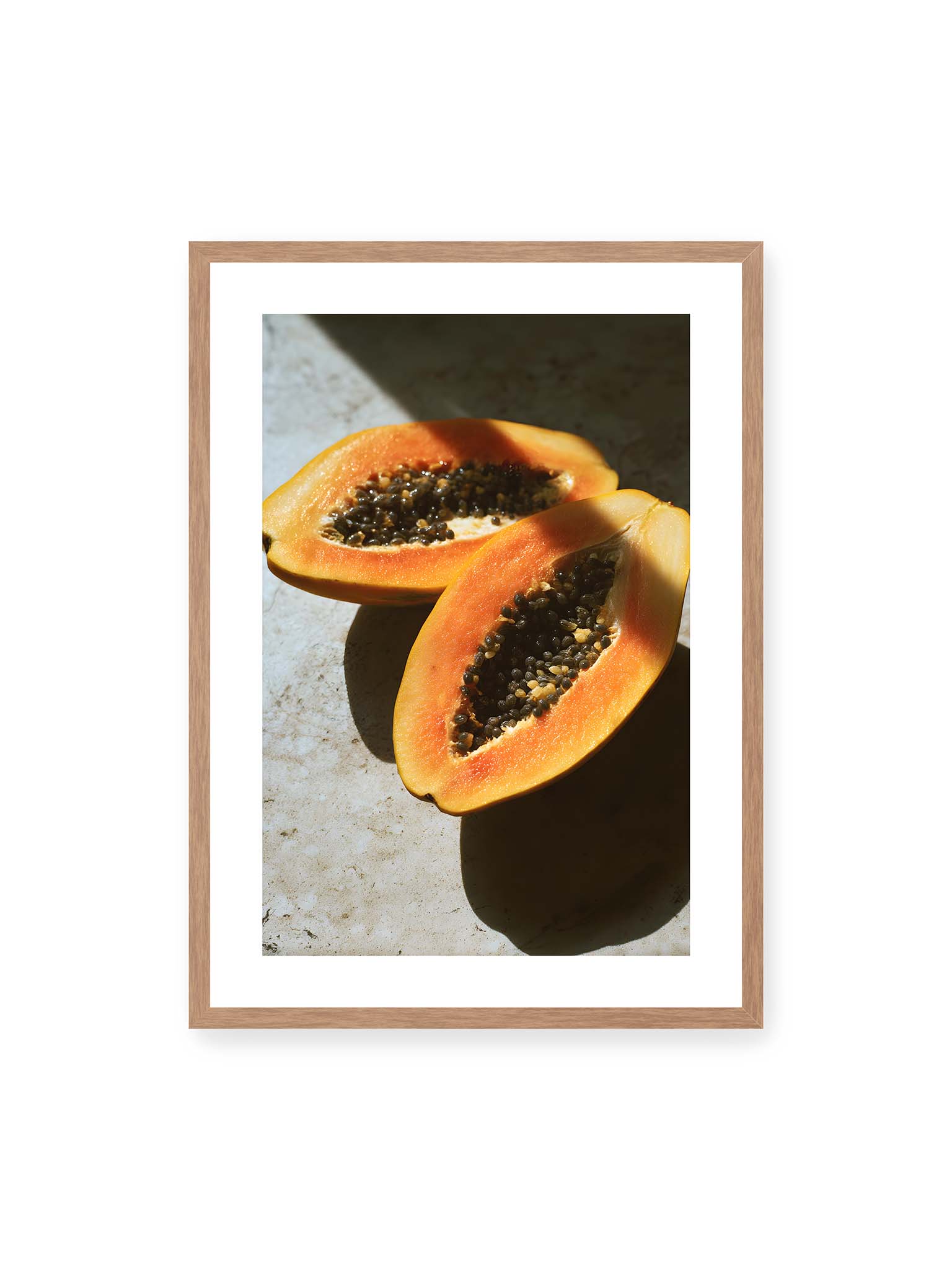 50x70 cm, weißer Galerie-Rand, Eichenrahmen – Zwei halbierte Papayas mit dunklen Samen auf heller Oberfläche, beleuchtet von warmem Licht mit starken Schatten.