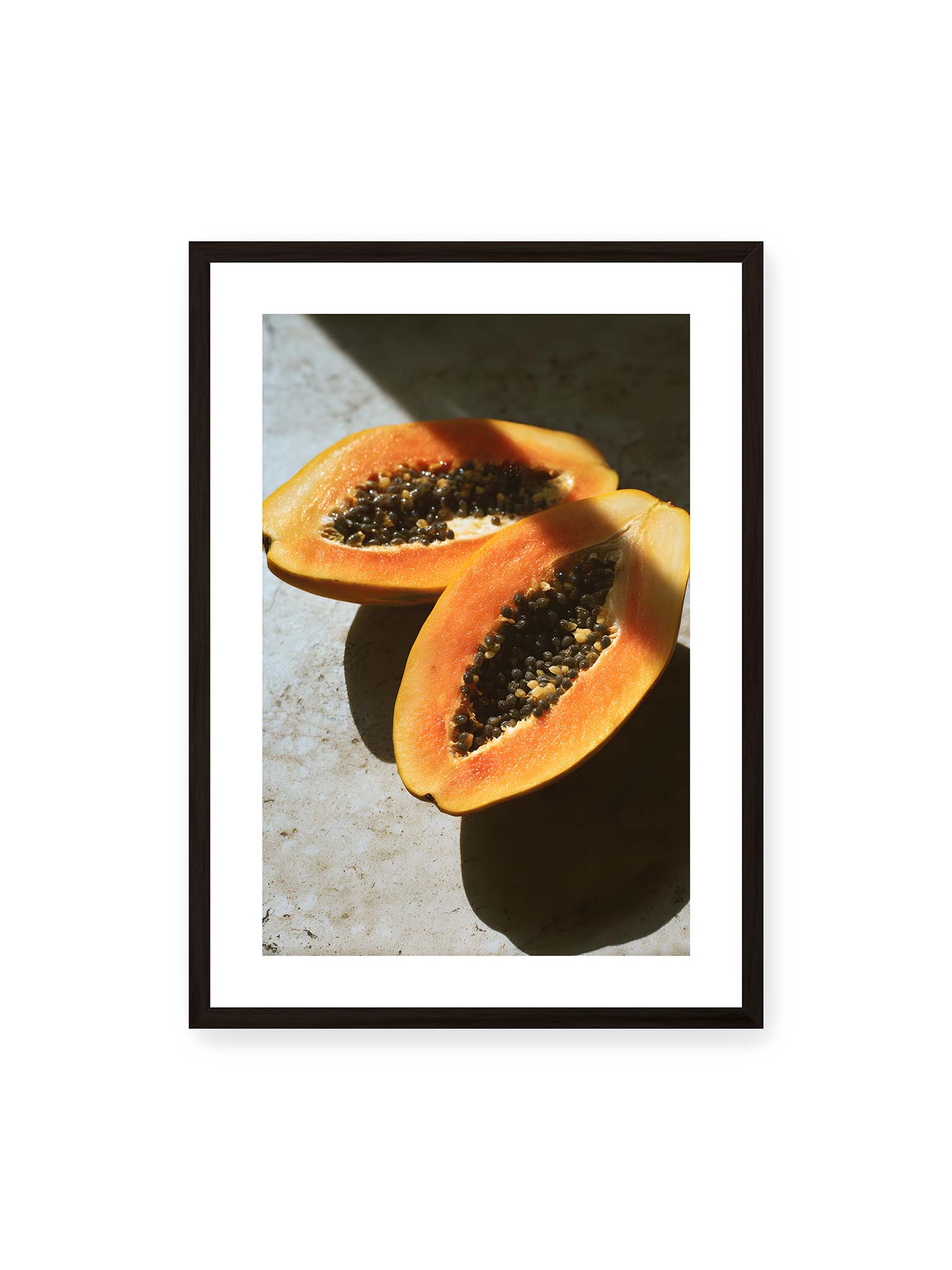 50x70 cm, weißer Galerie-Rand, schwarzer Rahmen – Zwei halbierte Papayas mit dunklen Samen auf heller Oberfläche, beleuchtet von warmem Licht mit starken Schatten.