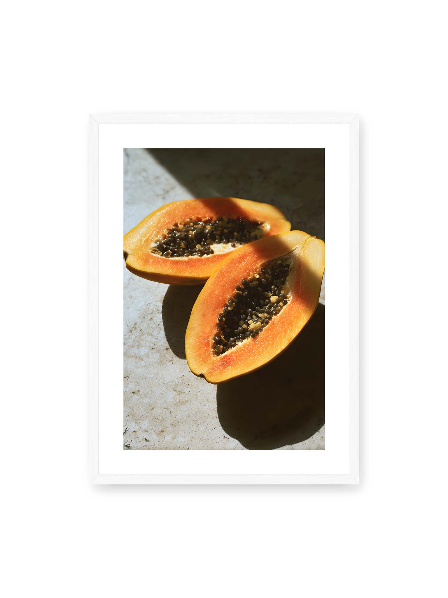 50x70 cm, weißer Galerie-Rand, weißer Rahmen – Zwei halbierte Papayas mit dunklen Samen auf heller Oberfläche, beleuchtet von warmem Licht mit starken Schatten.