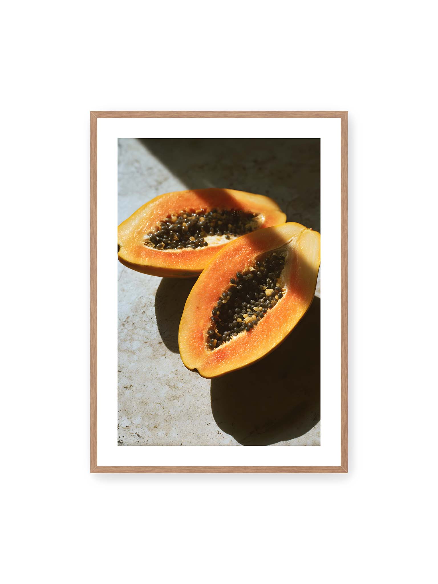 70x100 cm, weißer Galerie-Rand, Eichenrahmen – Zwei halbierte Papayas mit dunklen Samen auf heller Oberfläche, beleuchtet von warmem Licht mit starken Schatten.