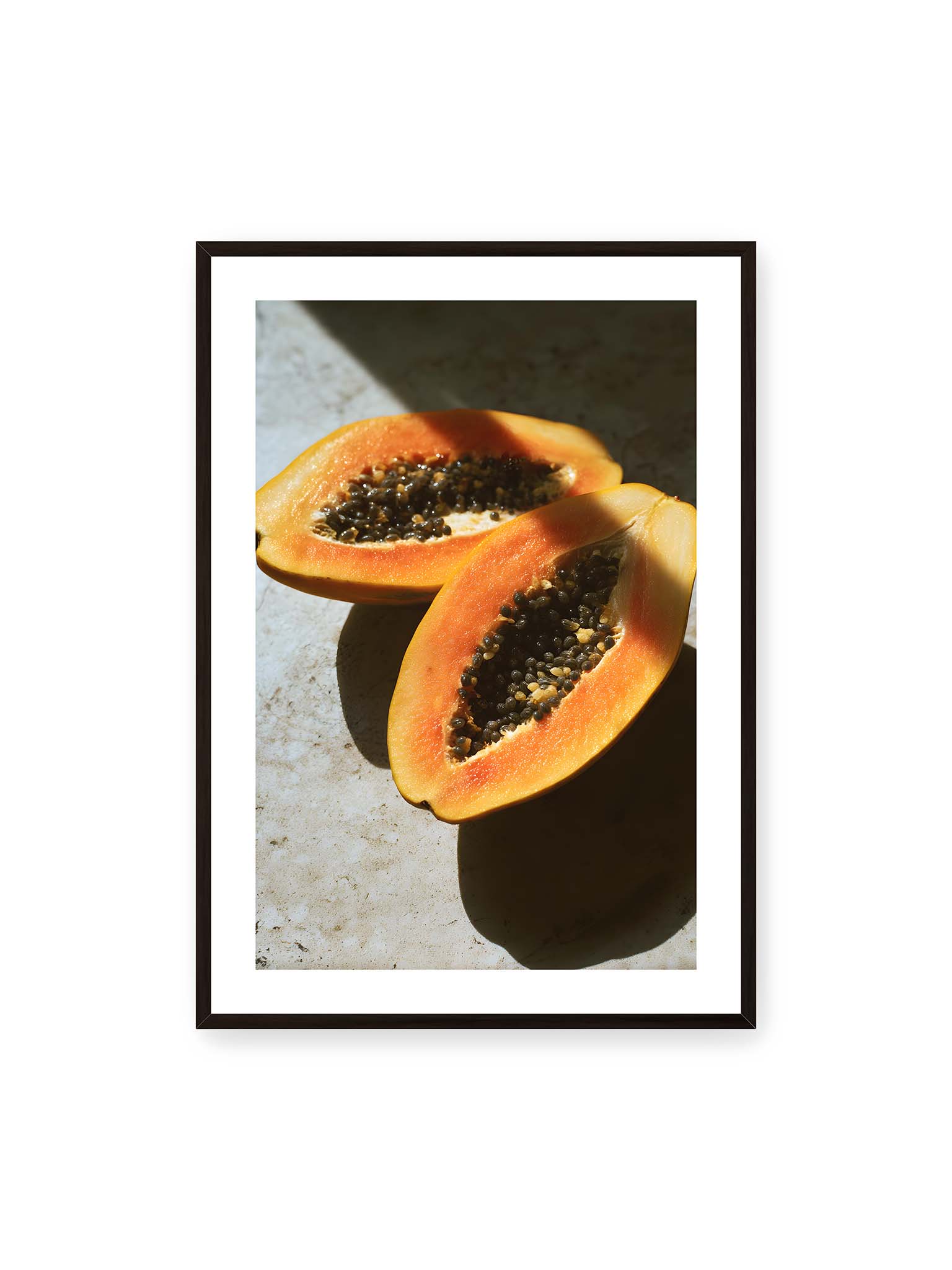 70x100 cm, weißer Galerie-Rand, schwarzer Rahmen – Zwei halbierte Papayas mit dunklen Samen auf heller Oberfläche, beleuchtet von warmem Licht mit starken Schatten.