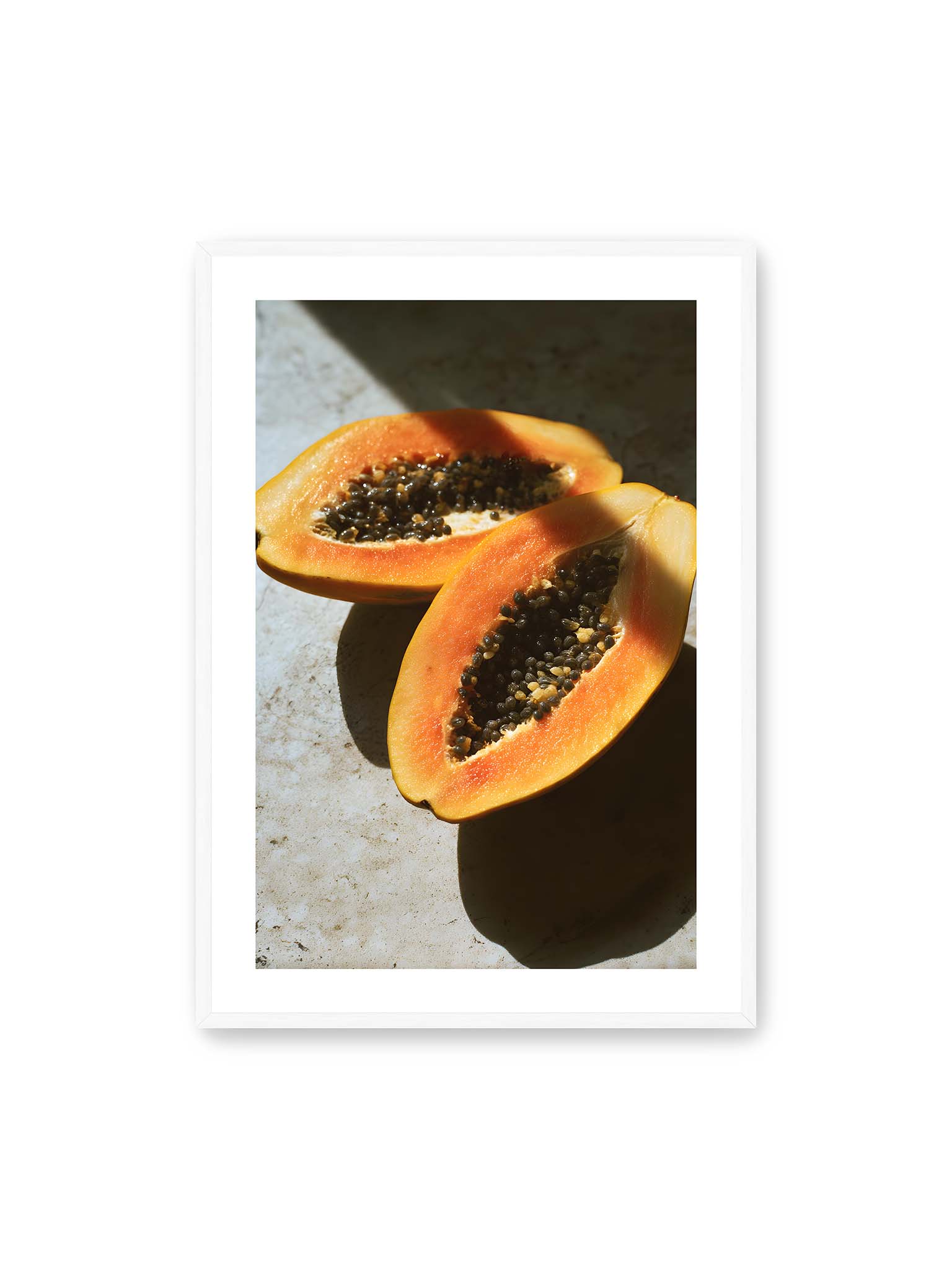 70x100 cm, weißer Galerie-Rand, weißer Rahmen – Zwei halbierte Papayas mit dunklen Samen auf heller Oberfläche, beleuchtet von warmem Licht mit starken Schatten.