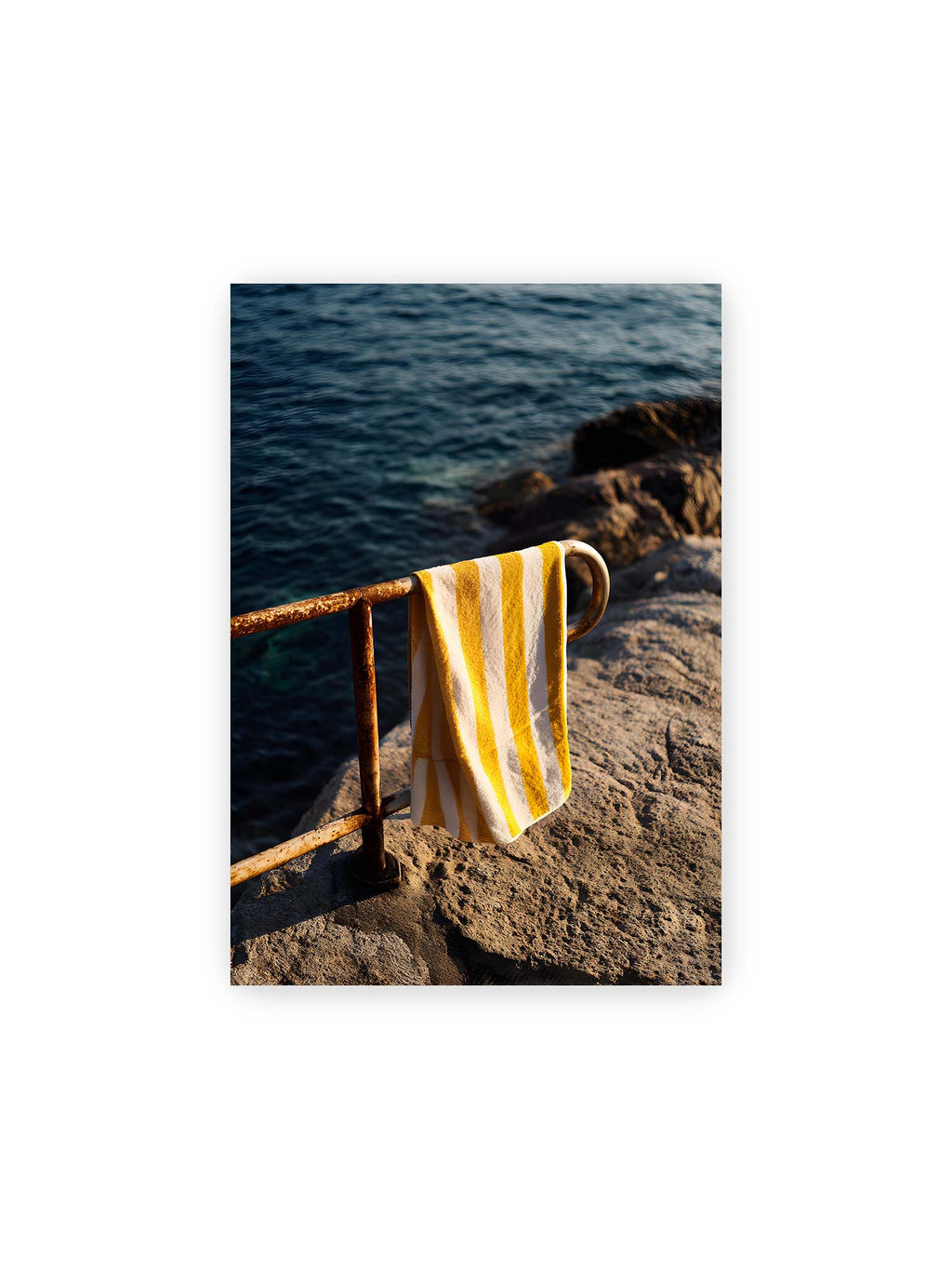 21x30 cm – Gelb-weiß gestreiftes Strandtuch hängt über rostigem Geländer am Meer bei warmem Licht.