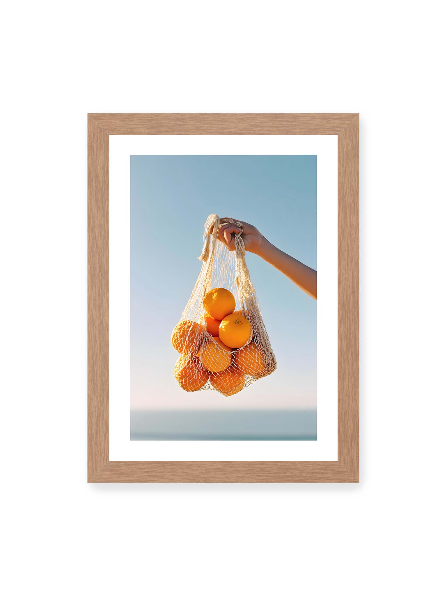21x30 cm, weißer Galerie-Rand, Eichenrahmen – Hand hält Netz mit Orangen vor klarem blauem Himmel mit starkem Farbkontrast.