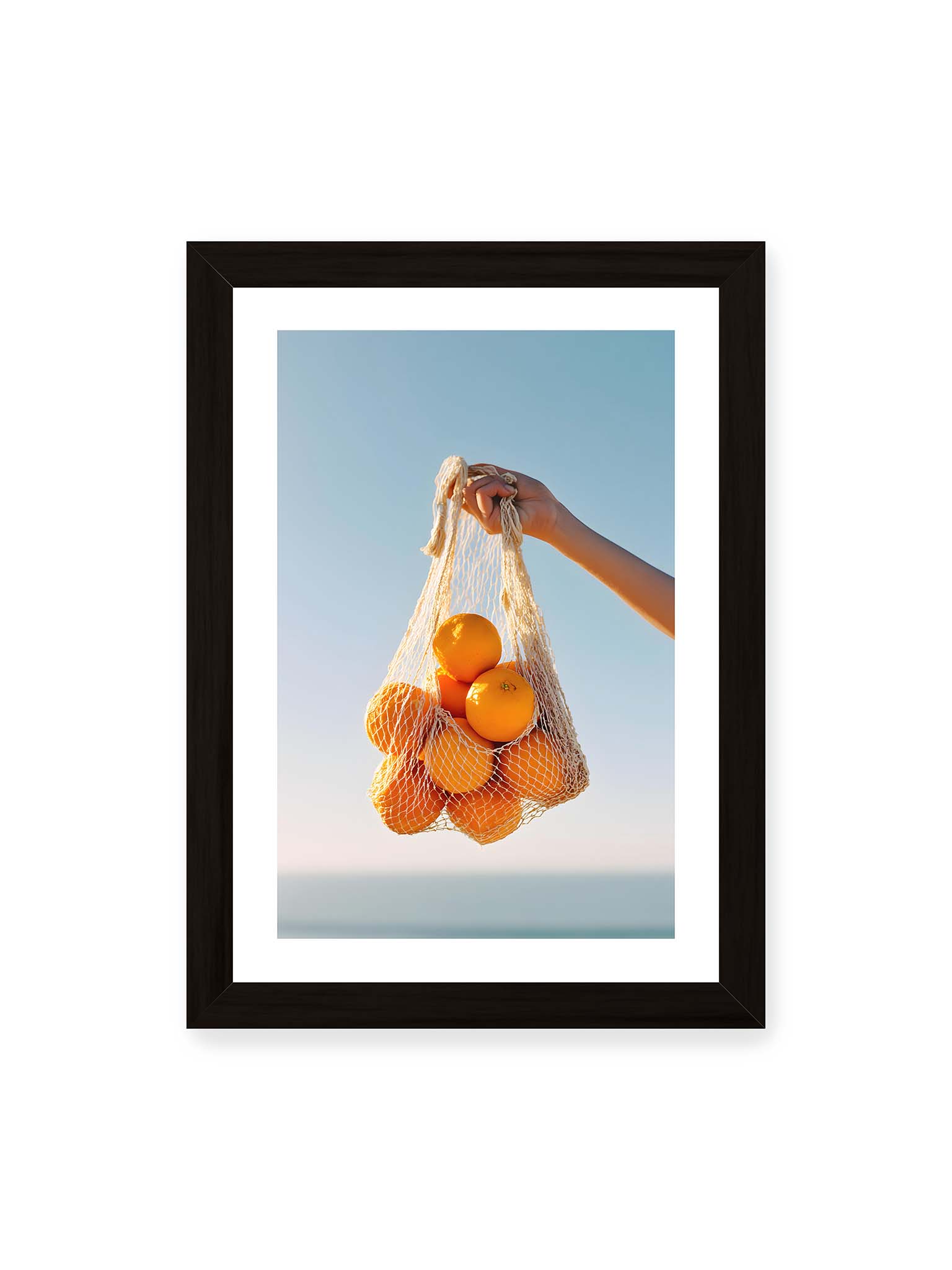 21x30 cm, weißer Galerie-Rand, schwarzer Rahmen – Hand hält Netz mit Orangen vor klarem blauem Himmel mit starkem Farbkontrast.