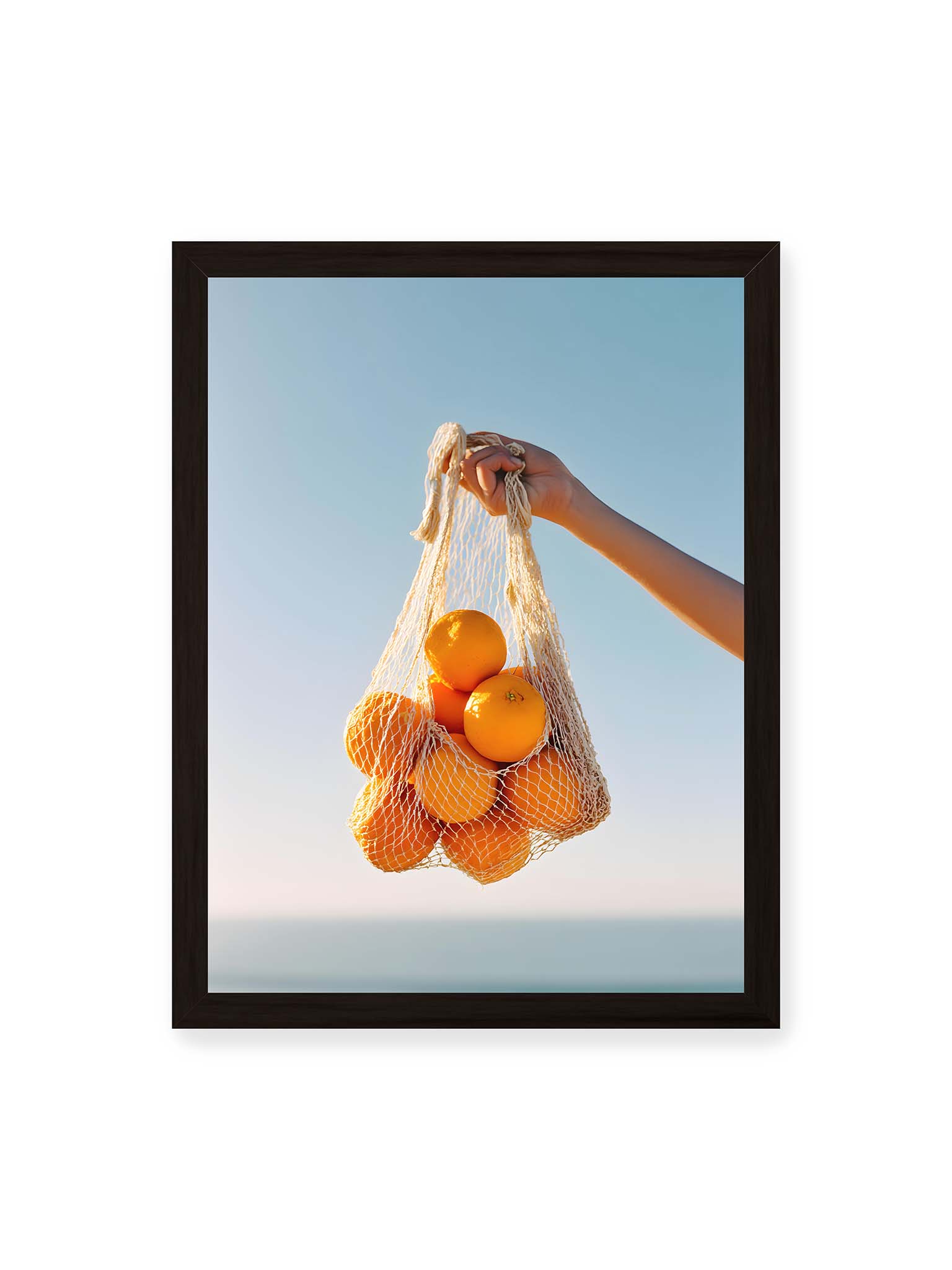 30x40 cm, schwarzer Rahmen – Hand hält Netz mit Orangen vor klarem blauem Himmel mit starkem Farbkontrast.