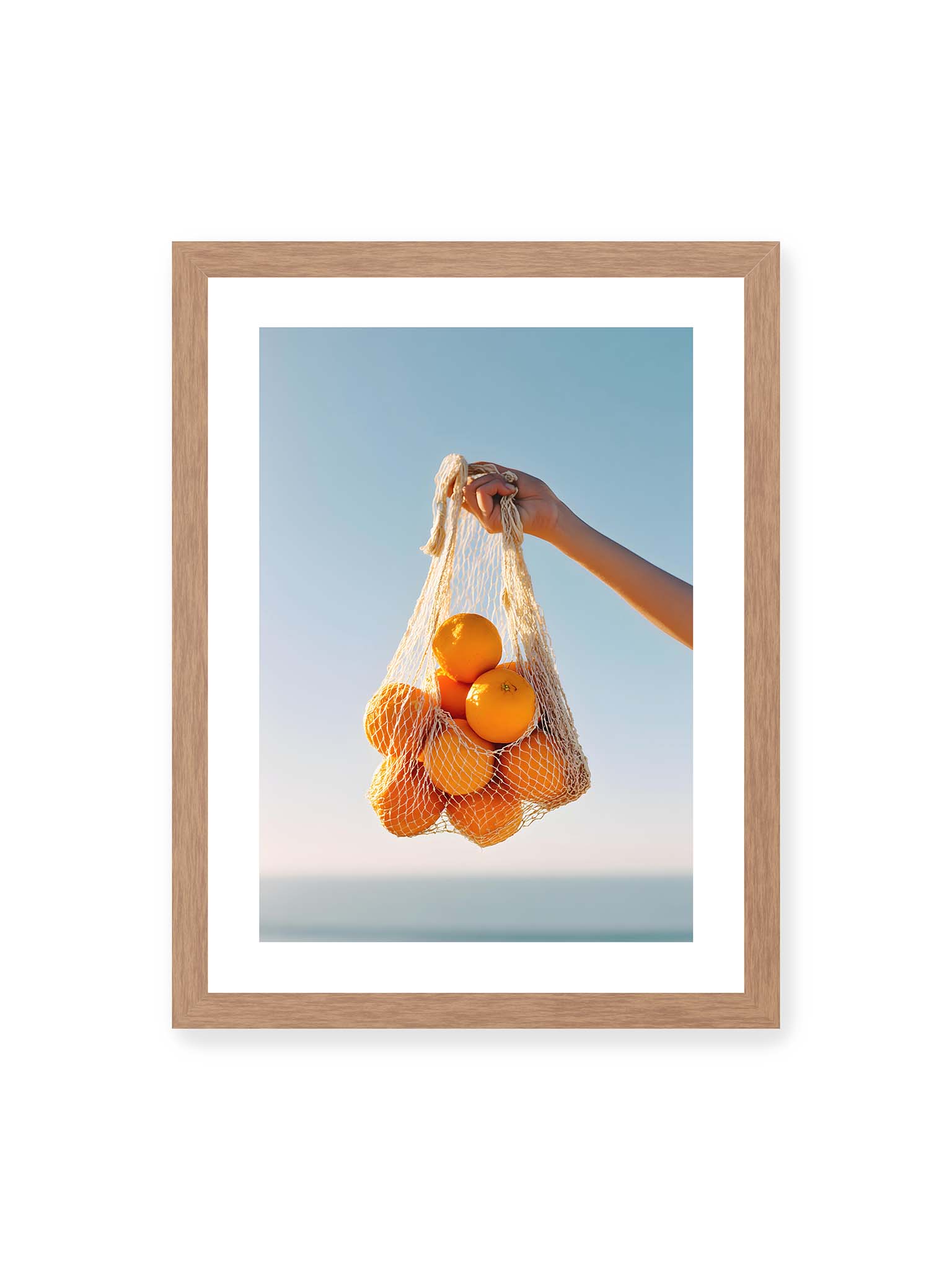 30x40 cm, weißer Galerie-Rand, Eichenrahmen – Hand hält Netz mit Orangen vor klarem blauem Himmel mit starkem Farbkontrast.
