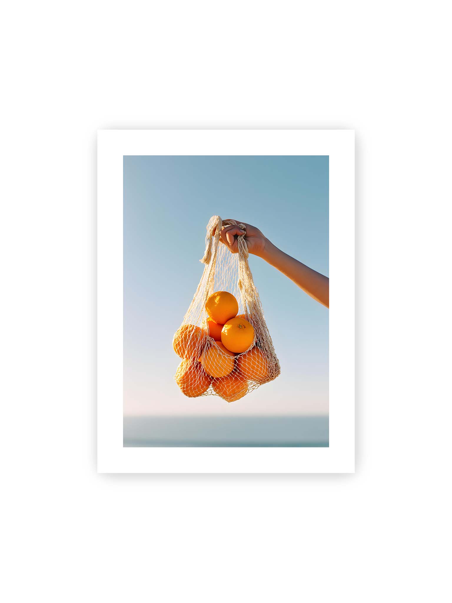 30x40 cm, weißer Galerie-Rand – Hand hält Netz mit Orangen vor klarem blauem Himmel mit starkem Farbkontrast.