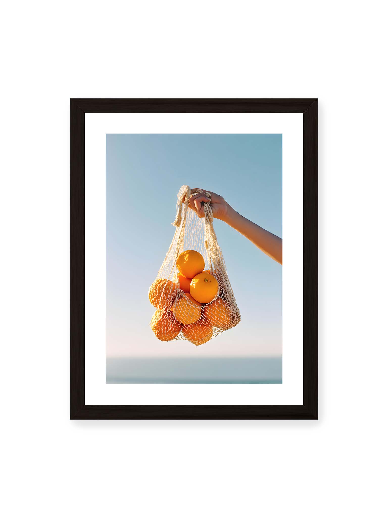 30x40 cm, weißer Galerie-Rand, schwarzer Rahmen – Hand hält Netz mit Orangen vor klarem blauem Himmel mit starkem Farbkontrast.