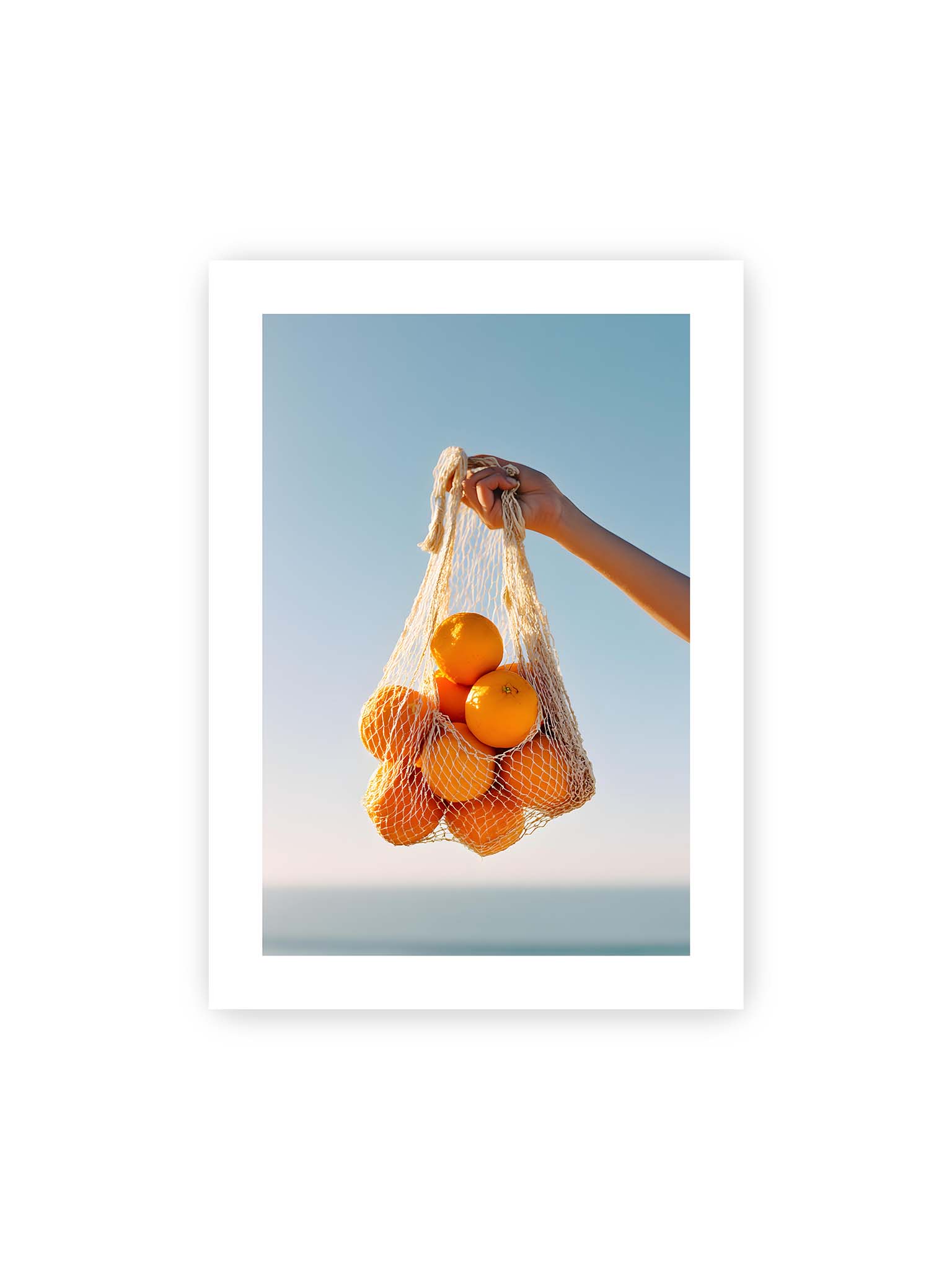 50x70 cm, weißer Galerie-Rand – Hand hält Netz mit Orangen vor klarem blauem Himmel mit starkem Farbkontrast.