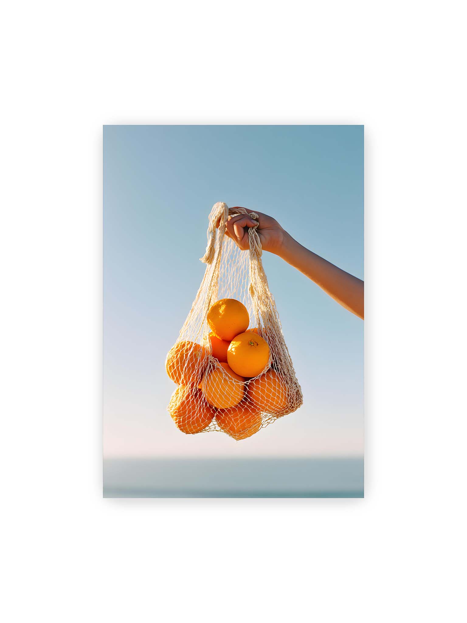 70x100 cm – Hand hält Netz mit Orangen vor klarem blauem Himmel mit starkem Farbkontrast.