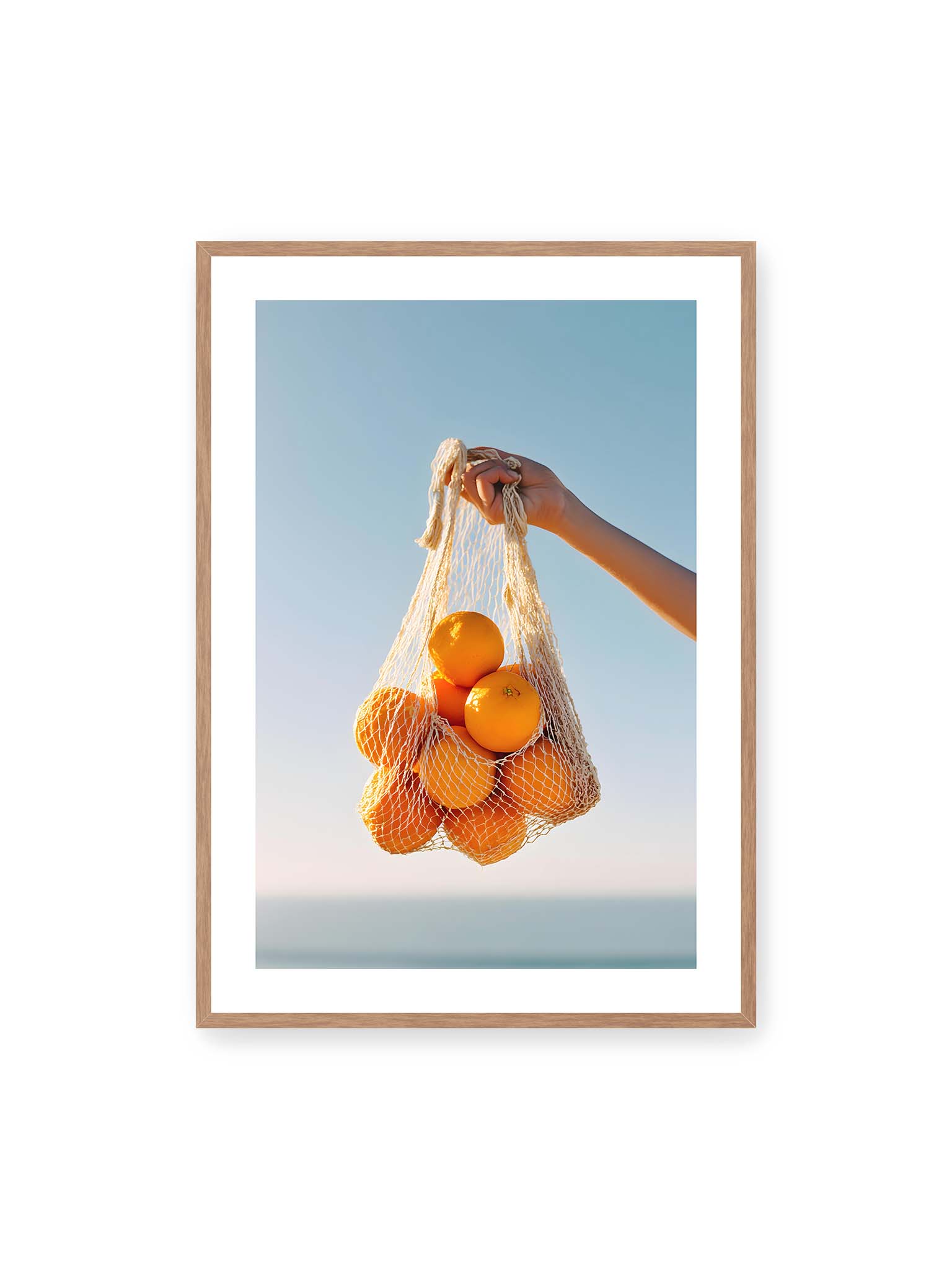 70x100 cm, weißer Galerie-Rand, Eichenrahmen – Hand hält Netz mit Orangen vor klarem blauem Himmel mit starkem Farbkontrast.