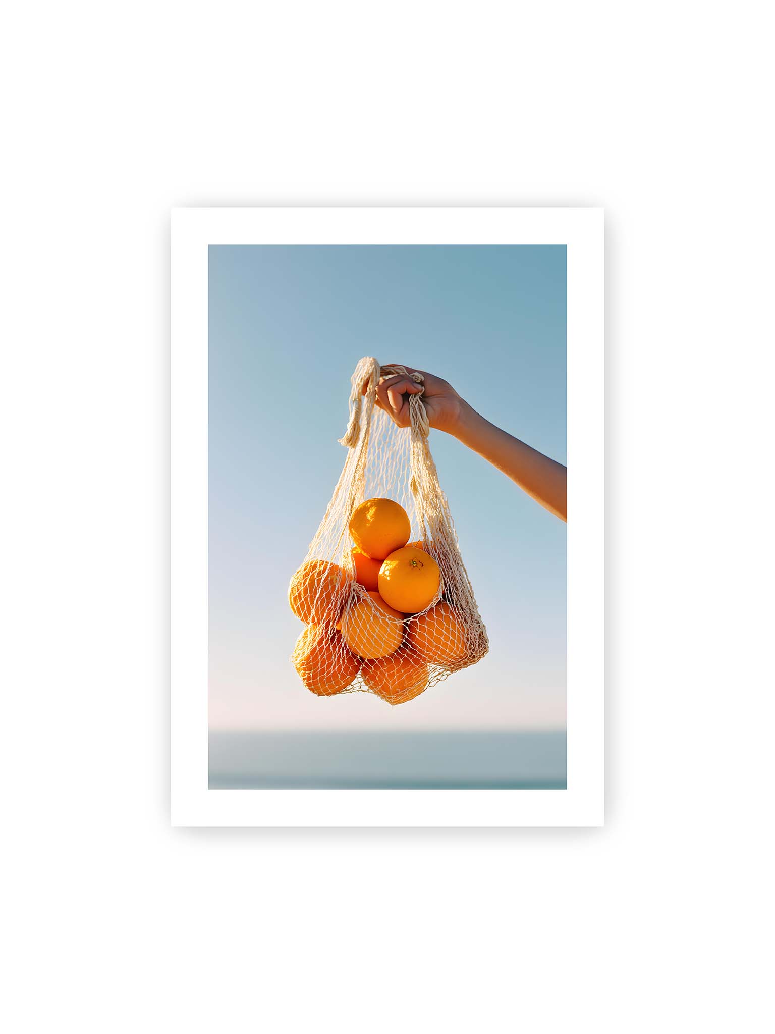 70x100 cm, weißer Galerie-Rand – Hand hält Netz mit Orangen vor klarem blauem Himmel mit starkem Farbkontrast.