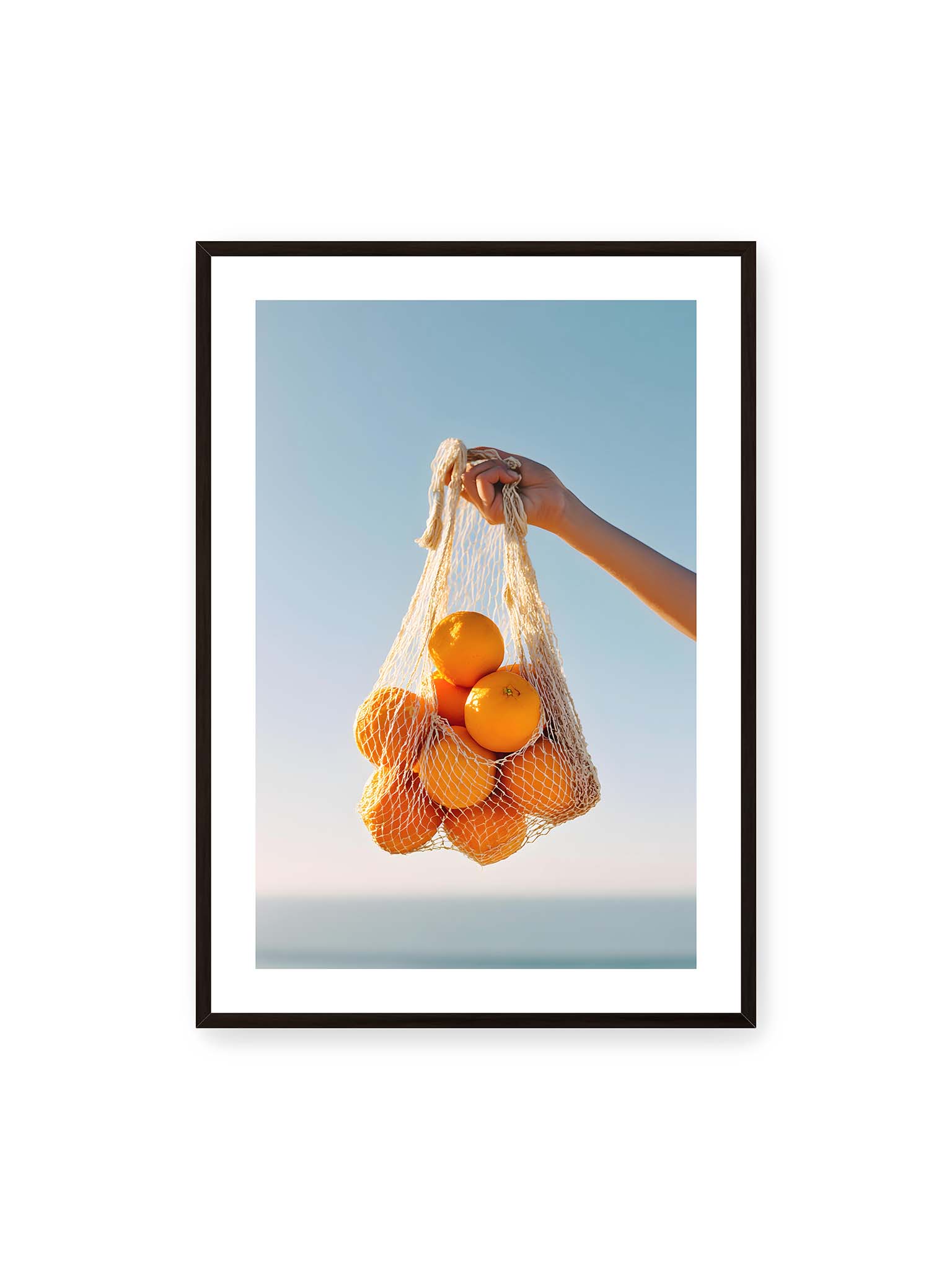 70x100 cm, weißer Galerie-Rand, schwarzer Rahmen – Hand hält Netz mit Orangen vor klarem blauem Himmel mit starkem Farbkontrast.