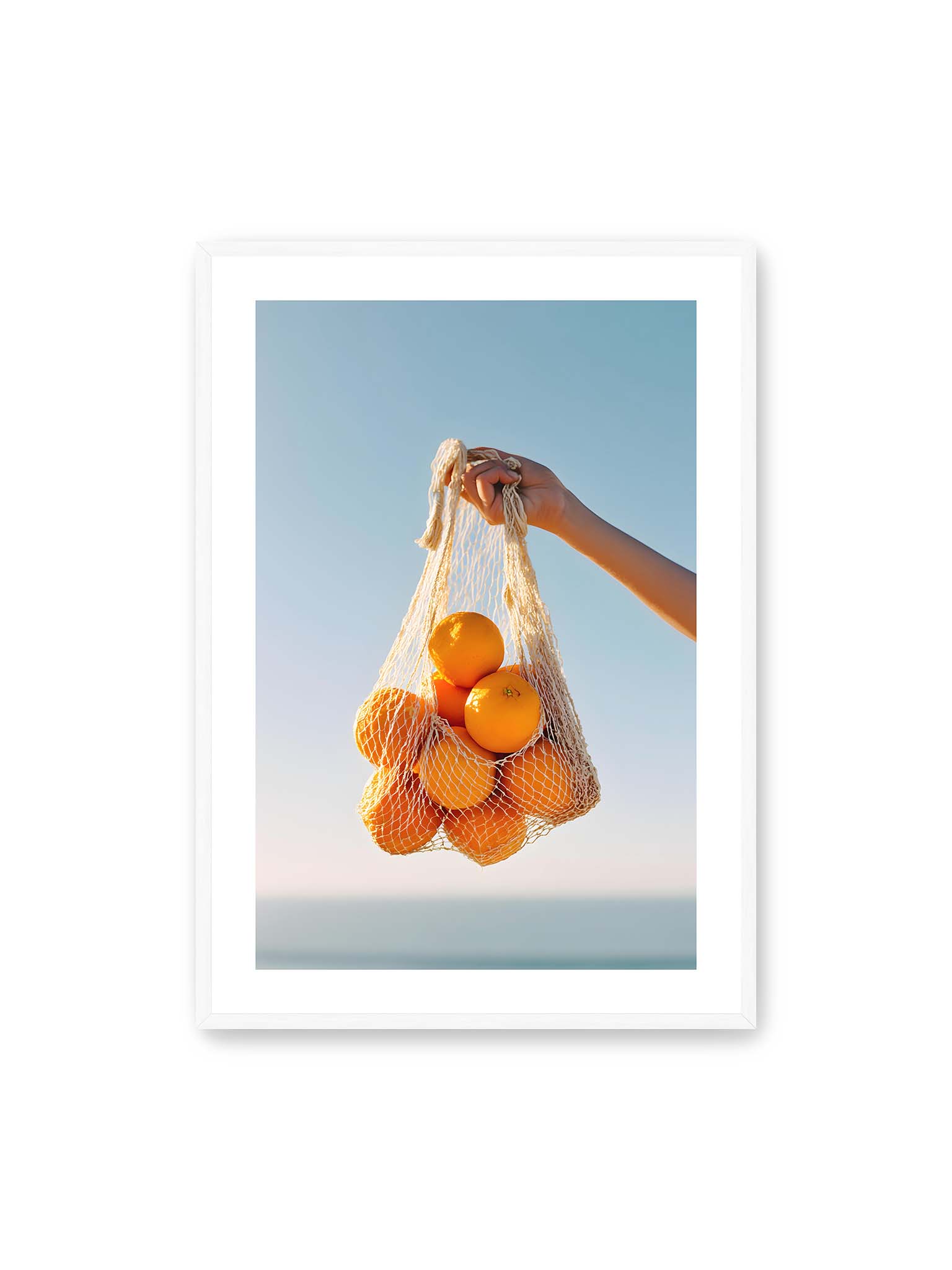 70x100 cm, weißer Galerie-Rand, weißer Rahmen – Hand hält Netz mit Orangen vor klarem blauem Himmel mit starkem Farbkontrast.