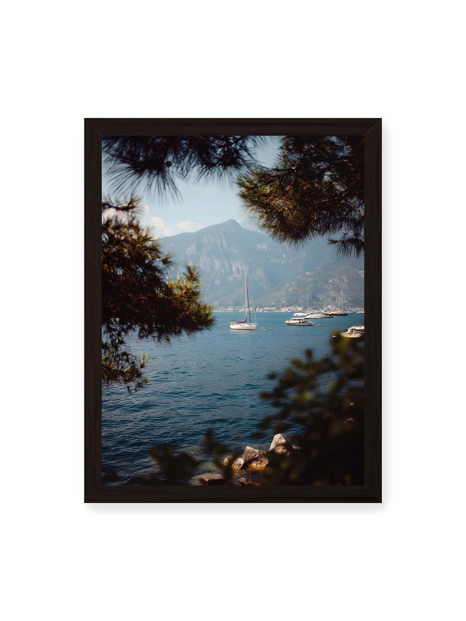 30x40 cm, schwarzer Rahmen – Segelboote im ruhigen Wasser vor Bergen, eingerahmt von unscharfen Ästen im Vordergrund.
