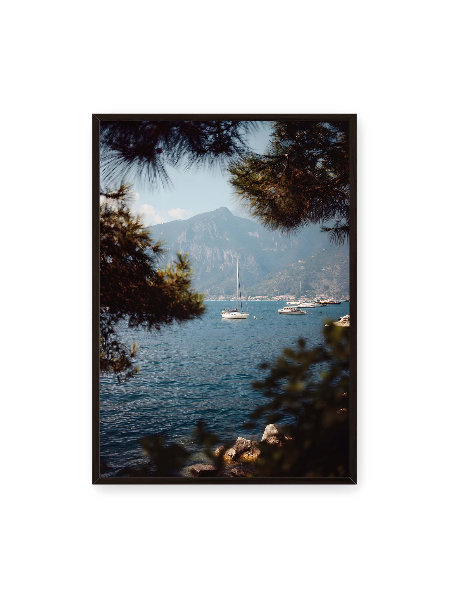 70x100 cm, schwarzer Rahmen – Segelboote im ruhigen Wasser vor Bergen, eingerahmt von unscharfen Ästen im Vordergrund.