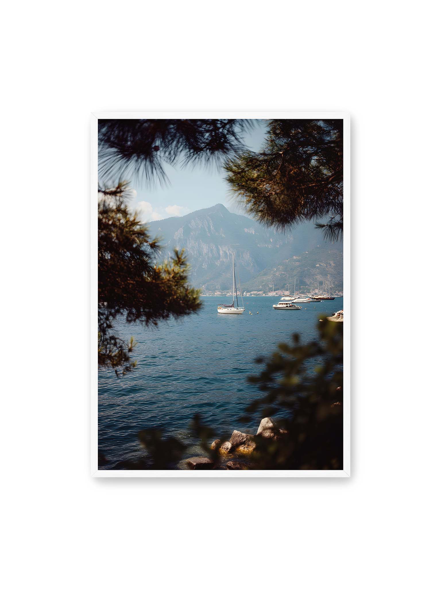70x100 cm, weißer Rahmen – Segelboote im ruhigen Wasser vor Bergen, eingerahmt von unscharfen Ästen im Vordergrund.