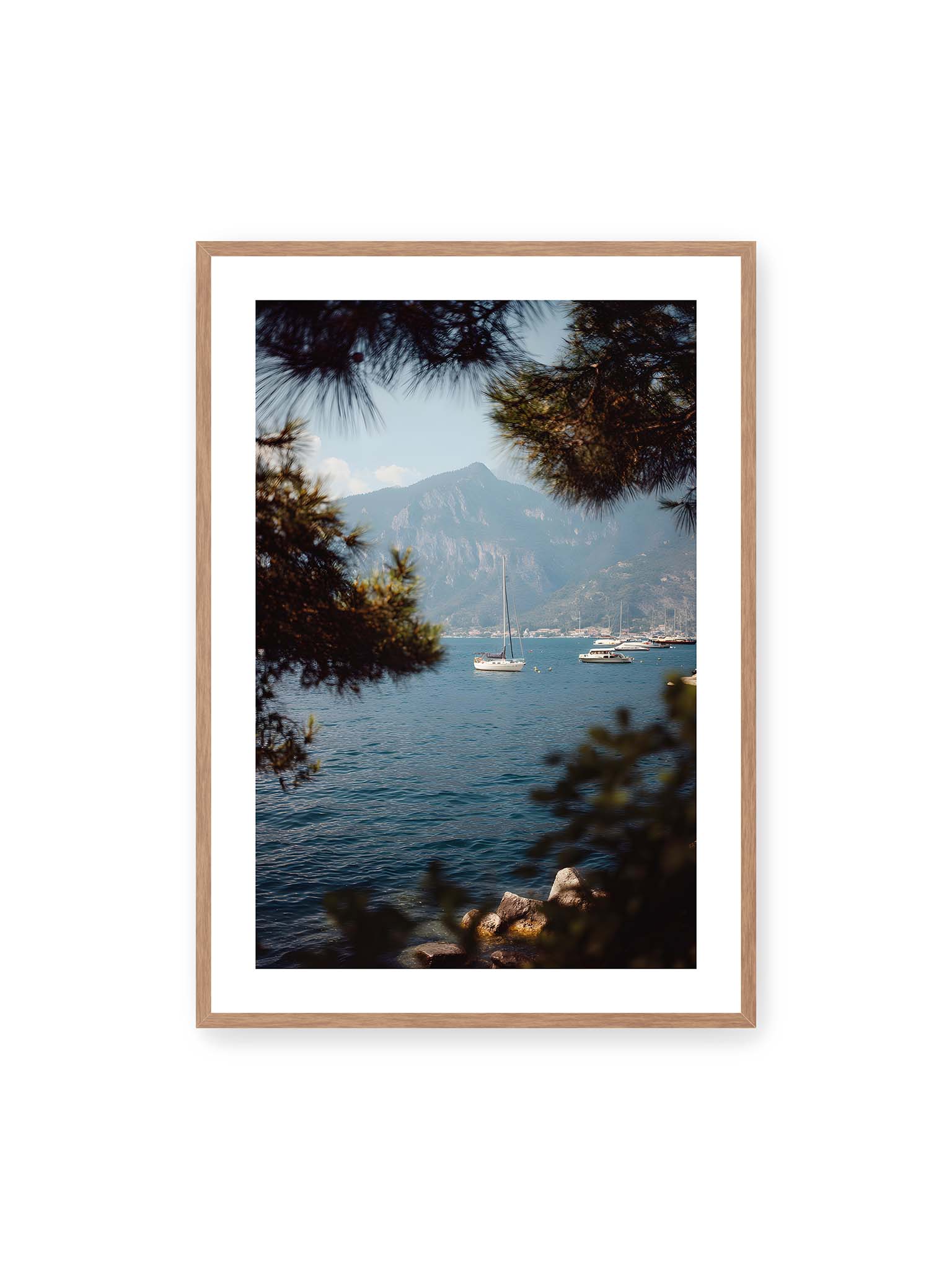 70x100 cm, weißer Galerie-Rand, Eichenrahmen – Segelboote im ruhigen Wasser vor Bergen, eingerahmt von unscharfen Ästen im Vordergrund.