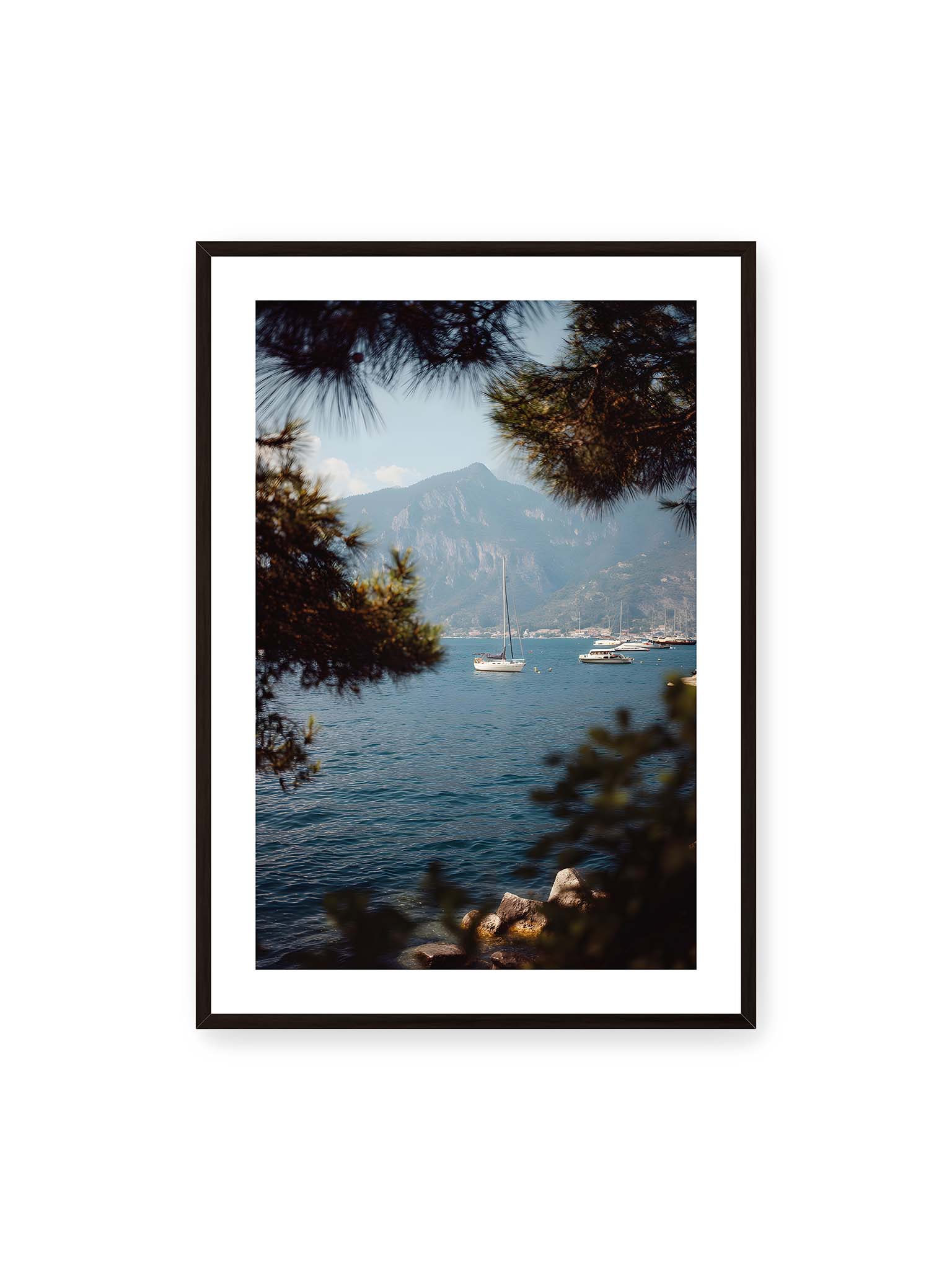 70x100 cm, weißer Galerie-Rand, schwarzer Rahmen – Segelboote im ruhigen Wasser vor Bergen, eingerahmt von unscharfen Ästen im Vordergrund.