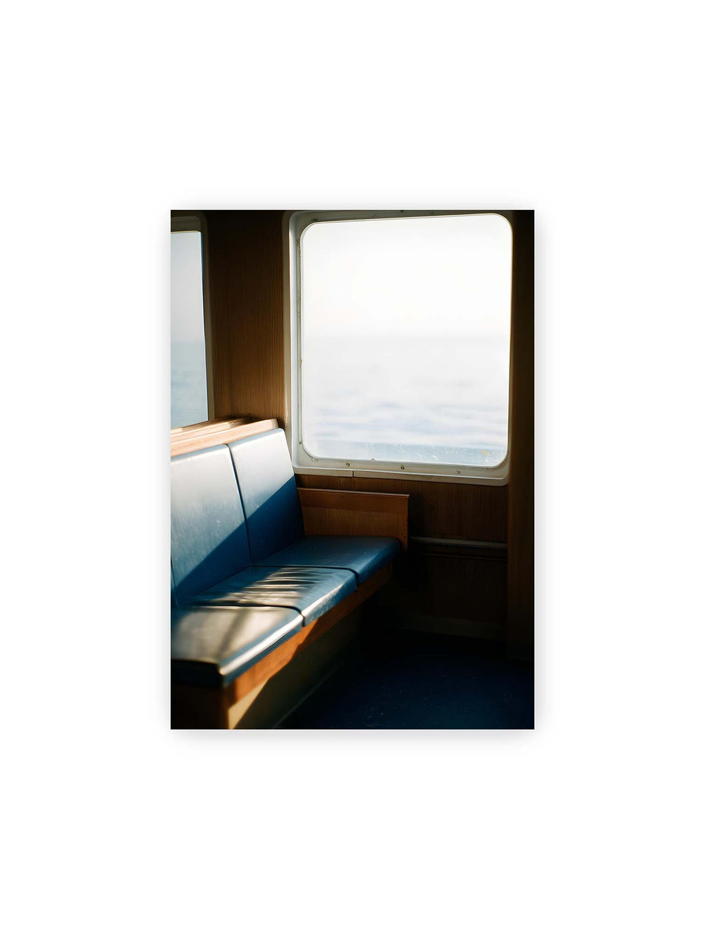 21x30 cm – Leerer Sitzplatz neben einem Fenster mit warmem Licht und unscharfer Aussicht auf das offene Meer.