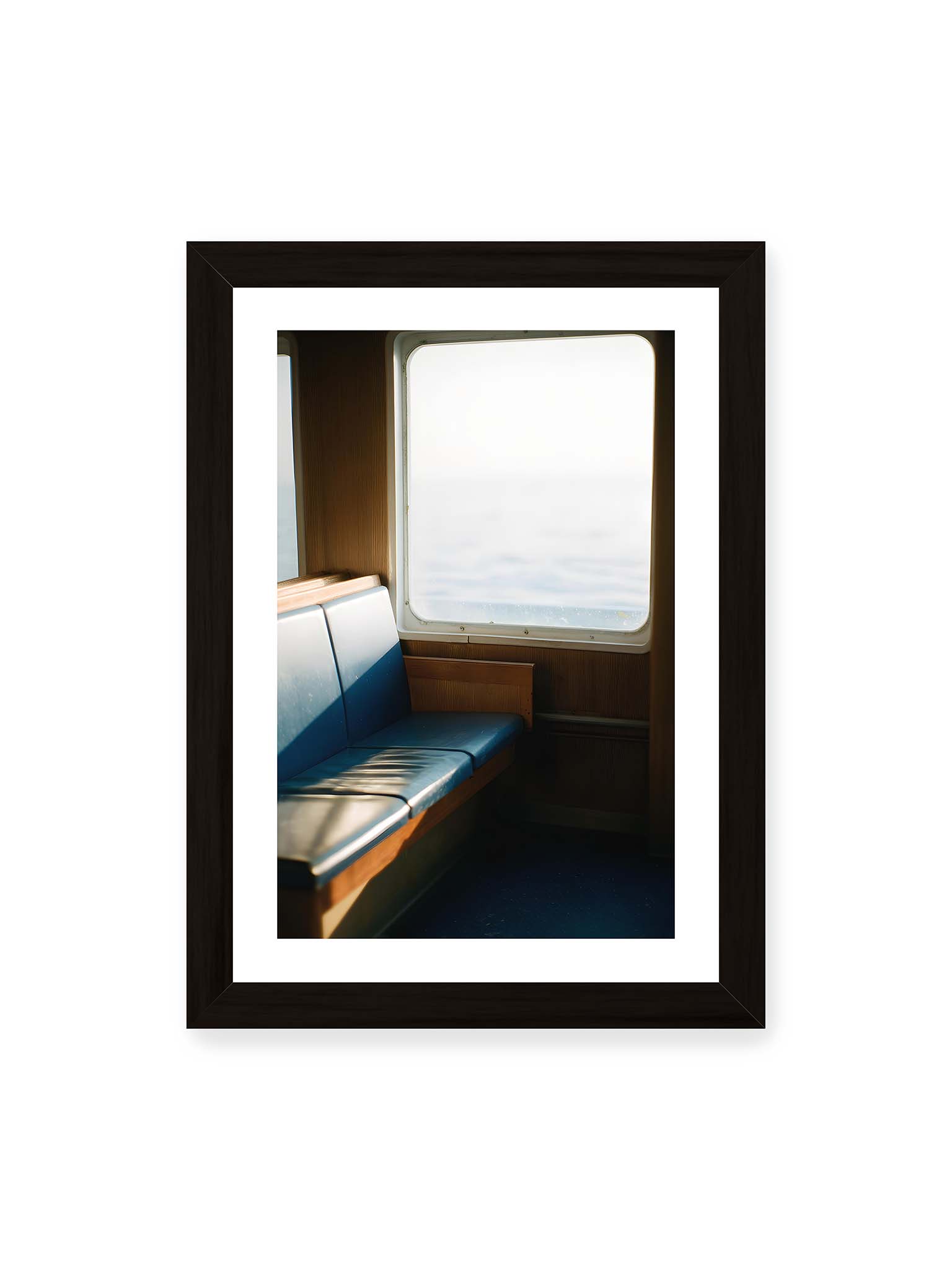 21x30 cm, weißer Galerie-Rand, schwarzer Rahmen – Leerer Sitzplatz neben einem Fenster mit warmem Licht und unscharfer Aussicht auf das offene Meer.