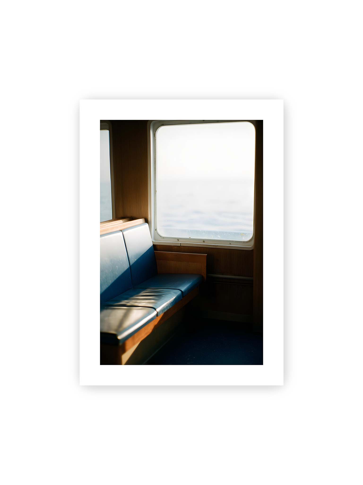 50x70 cm, weißer Galerie-Rand – Leerer Sitzplatz neben einem Fenster mit warmem Licht und unscharfer Aussicht auf das offene Meer.