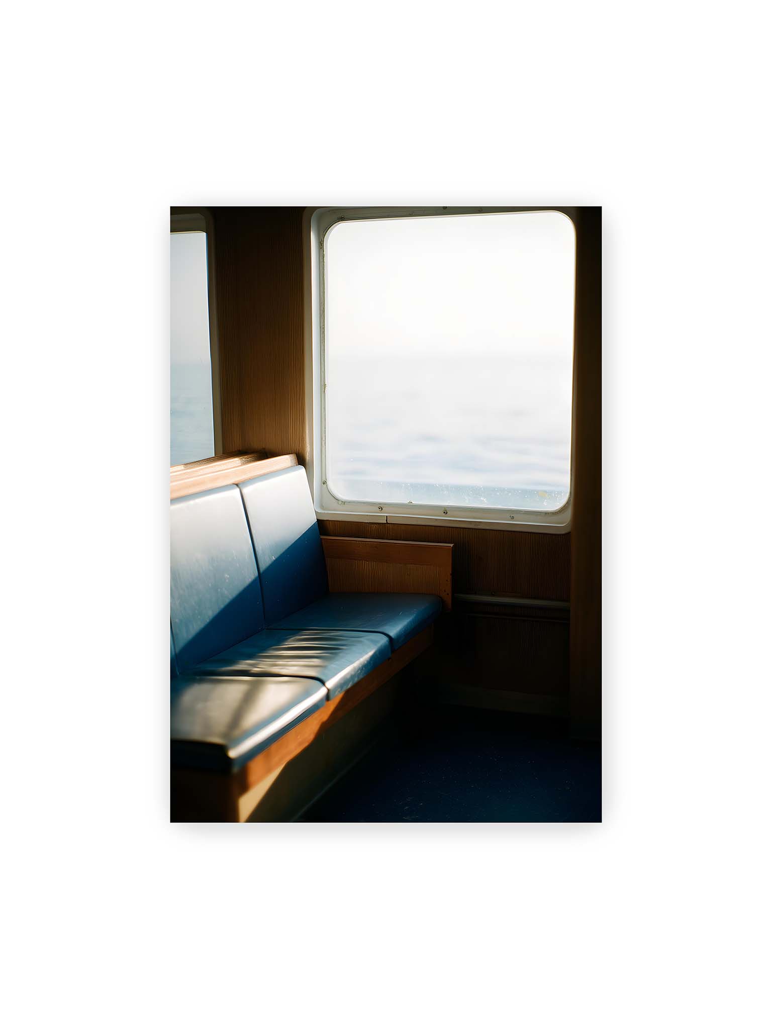 70x100 cm – Leerer Sitzplatz neben einem Fenster mit warmem Licht und unscharfer Aussicht auf das offene Meer.