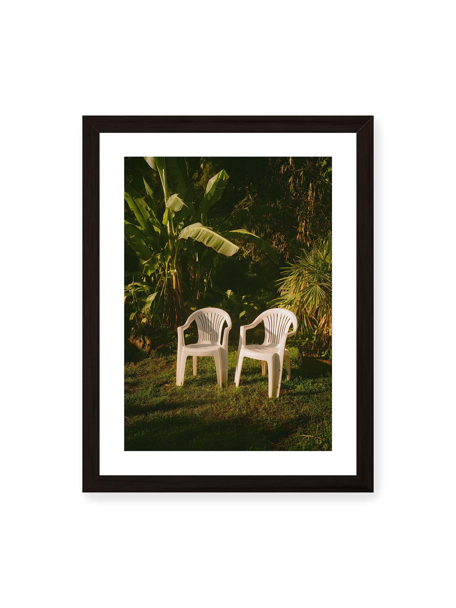 30x40 cm, weißer Galerie-Rand, schwarzer Rahmen – Zwei weiße Kunststoffstühle im tropischen Garten mit dichtem Grün und warmem Licht im Schattenbereich.