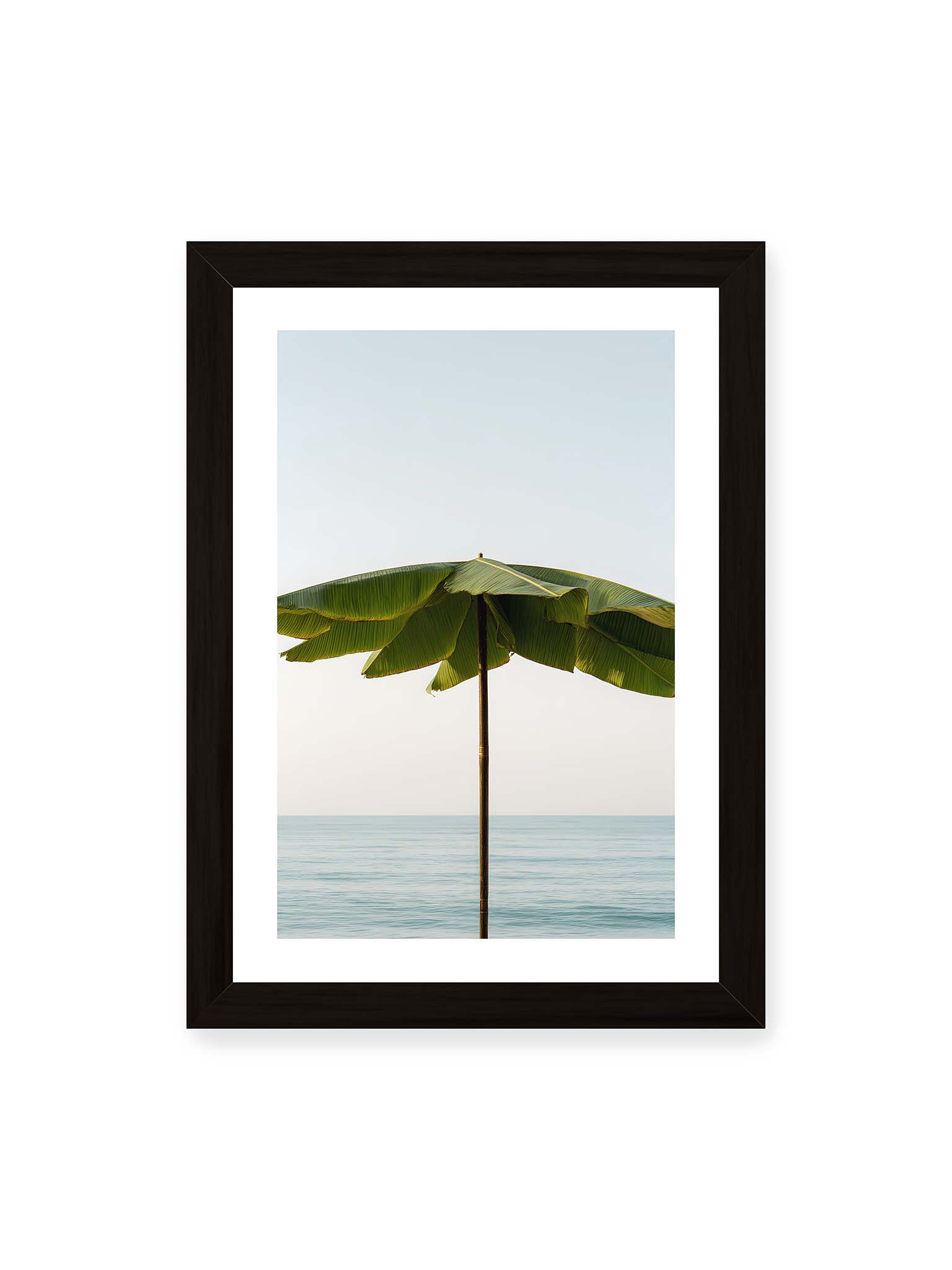 21x30 cm, weißer Galerie-Rand, schwarzer Rahmen – Sonnenschirm aus großen grünen tropischen Blättern vor ruhigem Meer mit klarer Horizontlinie und hellem Himmel.