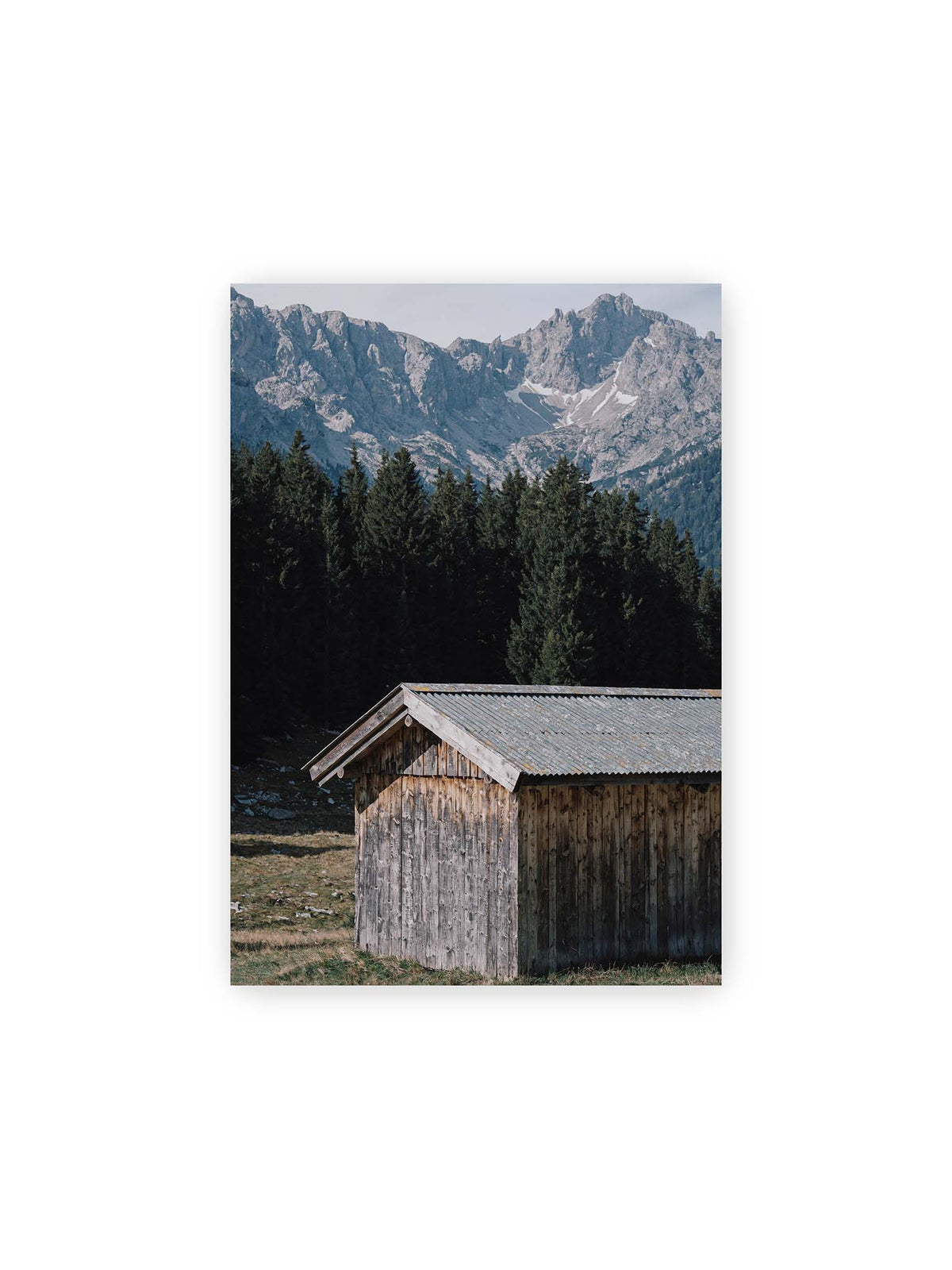 21x30 cm – Verwitterte Holzhütte vor dunklen Nadelbäumen und einer hellen, zerklüfteten Bergkulisse.